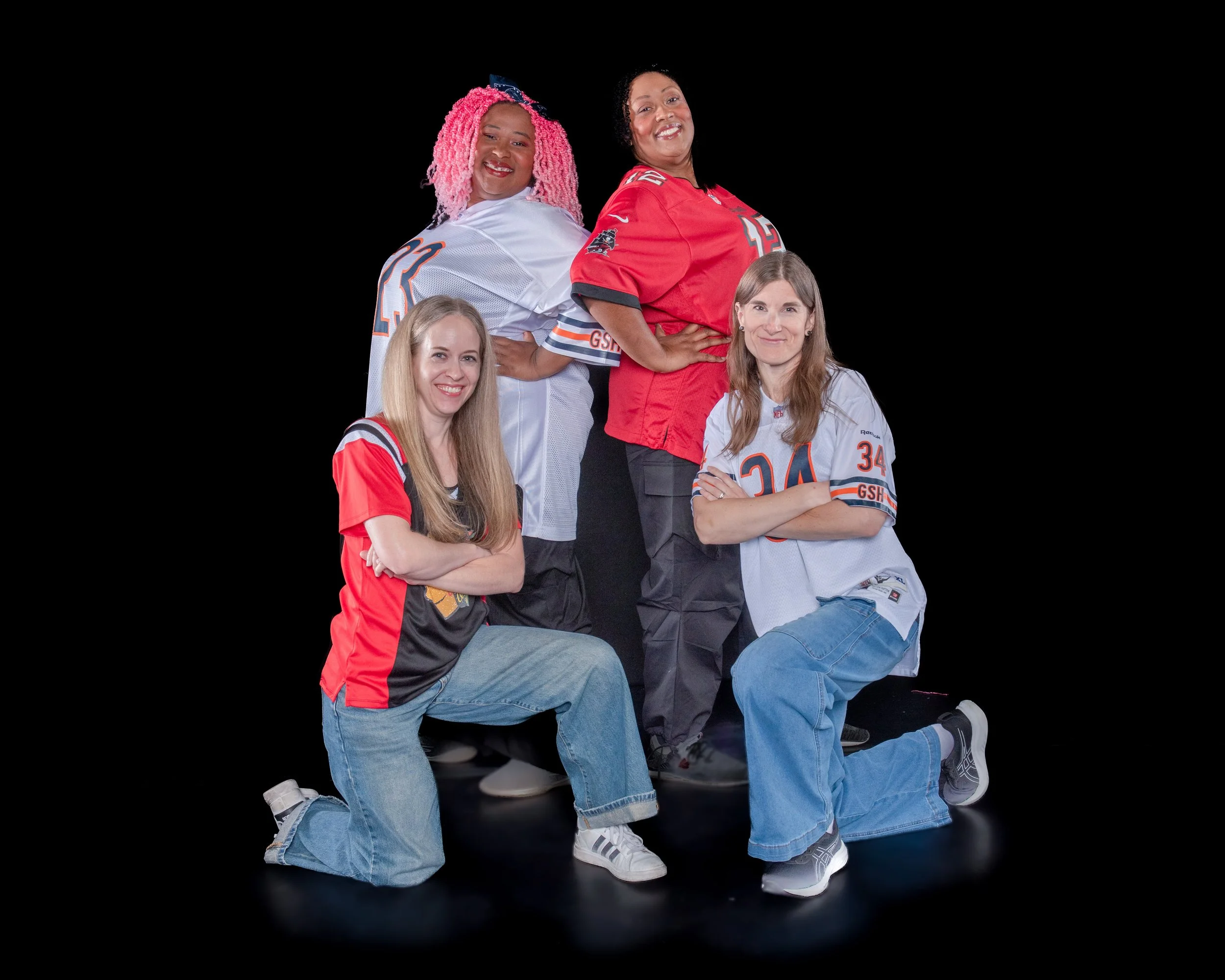 Group of four women posing together on a black background, wearing sports jerseys and casual clothing, smiling at the camera.