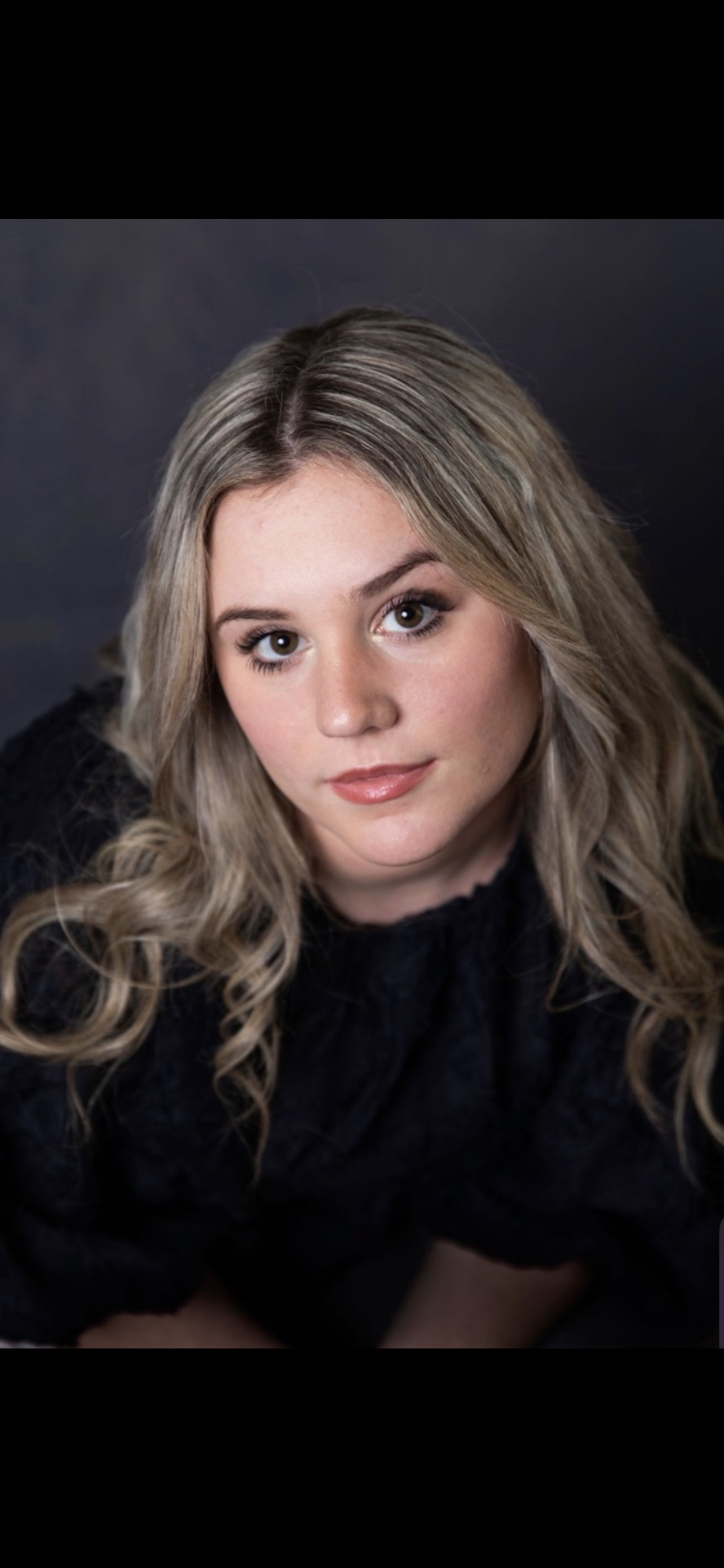 Portrait of a young woman with blonde, wavy hair and light makeup, wearing a black top, against a dark background.