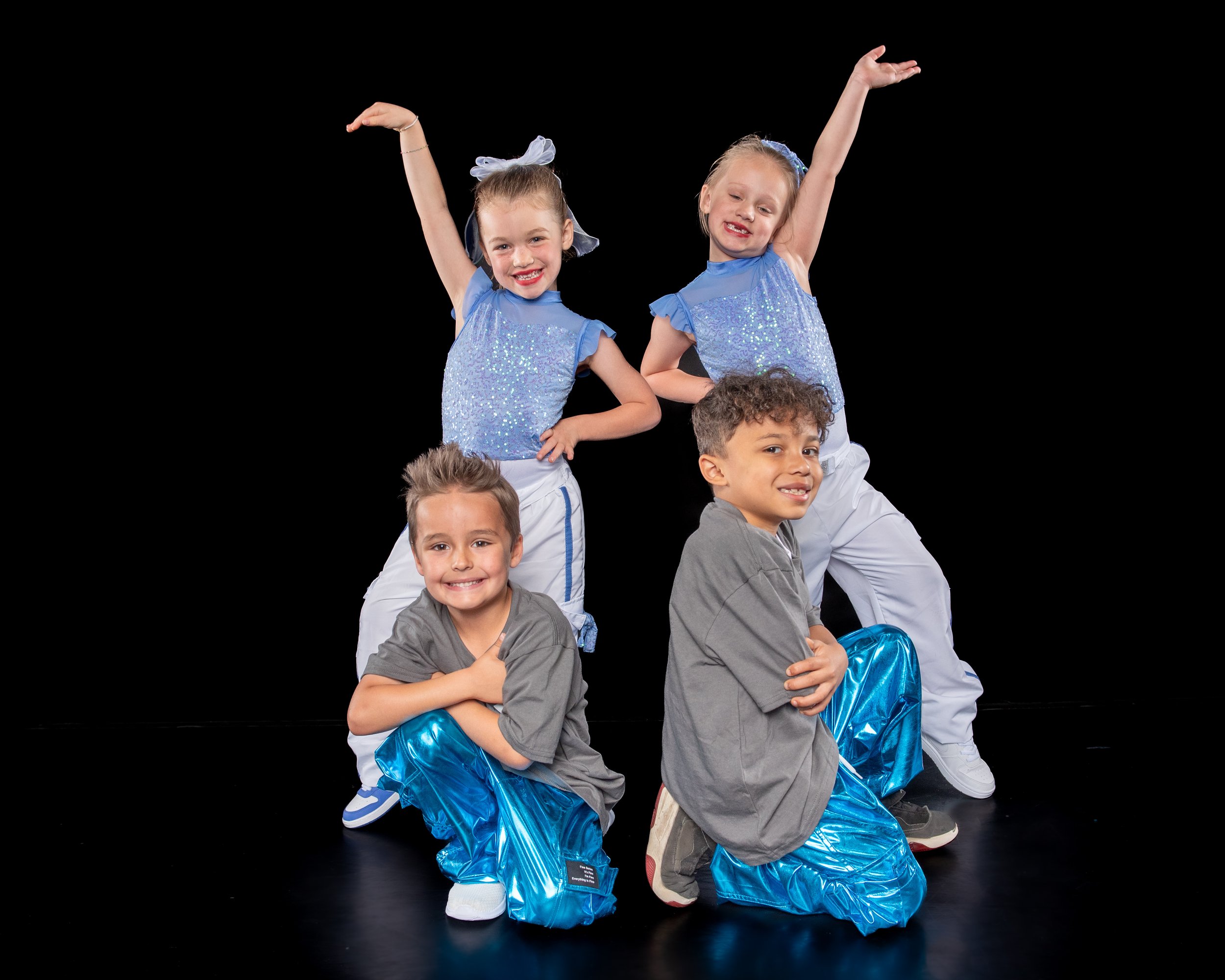 Four children in dance costumes, two girls in blue glittery tops and white pants, two boys in gray shirts and shiny blue pants, posing with arms raised and smiling against a black background.