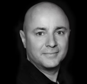 Black and white portrait of a bald man with light-colored eyes, smiling slightly, against a dark background.
