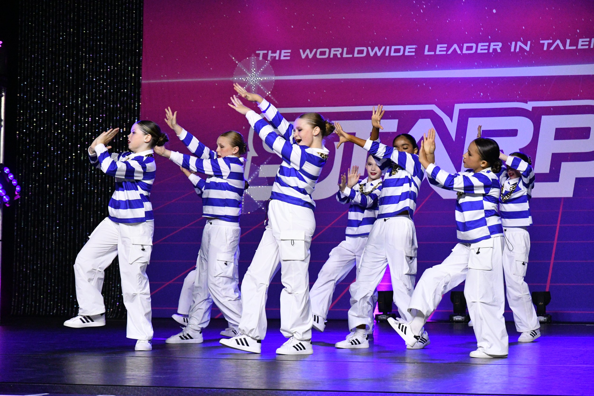 Group of young girls in matching blue and white striped shirts, white pants, and sneakers performing a choreographed dance on stage with a purple backdrop that says 'The Worldwide Leader in Talent' and 'GANE'. The girls are in synchronized poses with some raising their hands and others with hands near their faces.
