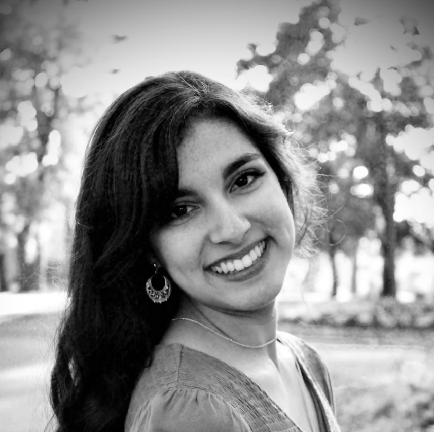 A smiling young woman with dark hair, wearing earrings and a necklace, outdoors with trees in the background.