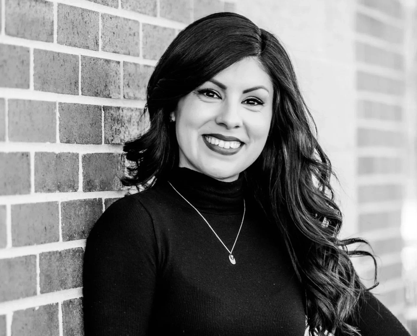 Black and white portrait of a woman with long wavy hair, smiling, wearing a turtleneck sweater and a necklace, leaning against a brick wall.