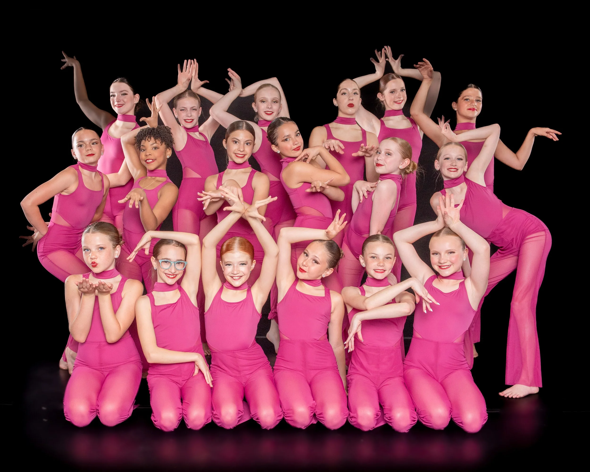 A group of young female dancers wearing matching pink costumes and posing against a black background.