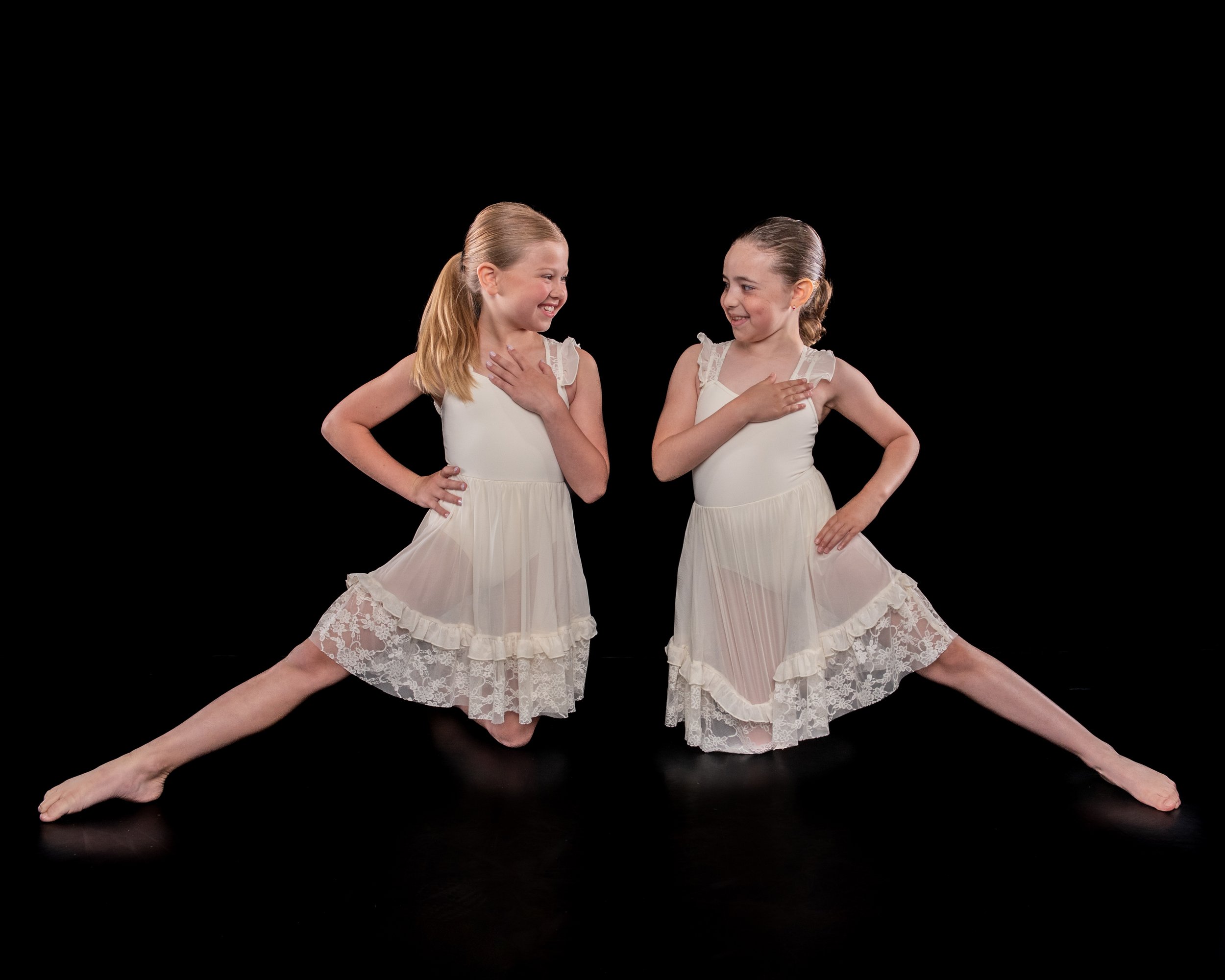 Two young girls dressed in white dance dresses practicing ballet, posing with one leg extended and hands on their chests, against a black background.
