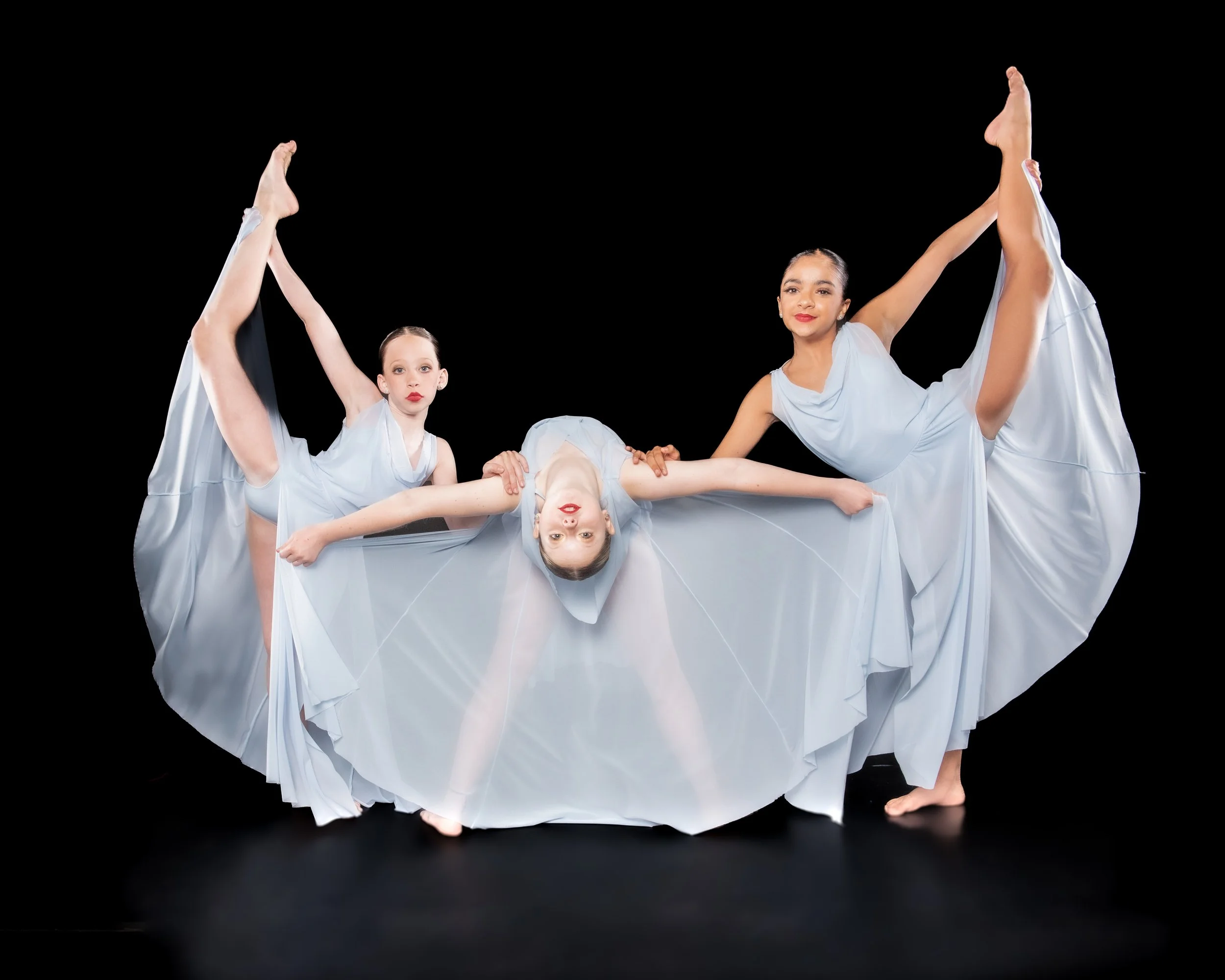 Two young female ballet dancers dressed in light blue costumes performing a pose against a black background, with their bodies intertwined and their arms extended.