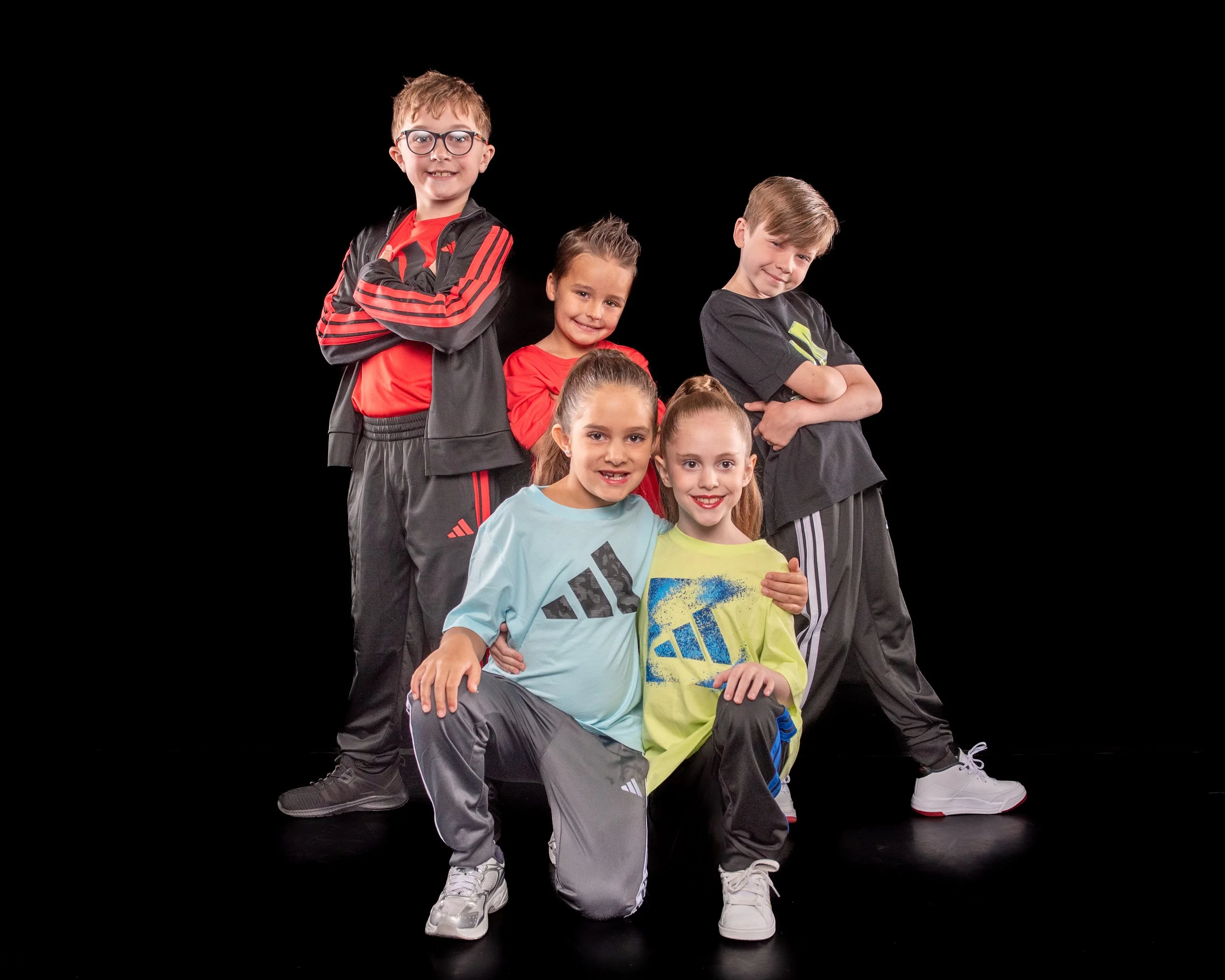 Group of six children posing together against a black background, some standing and some crouching, with crossed arms and smiling.