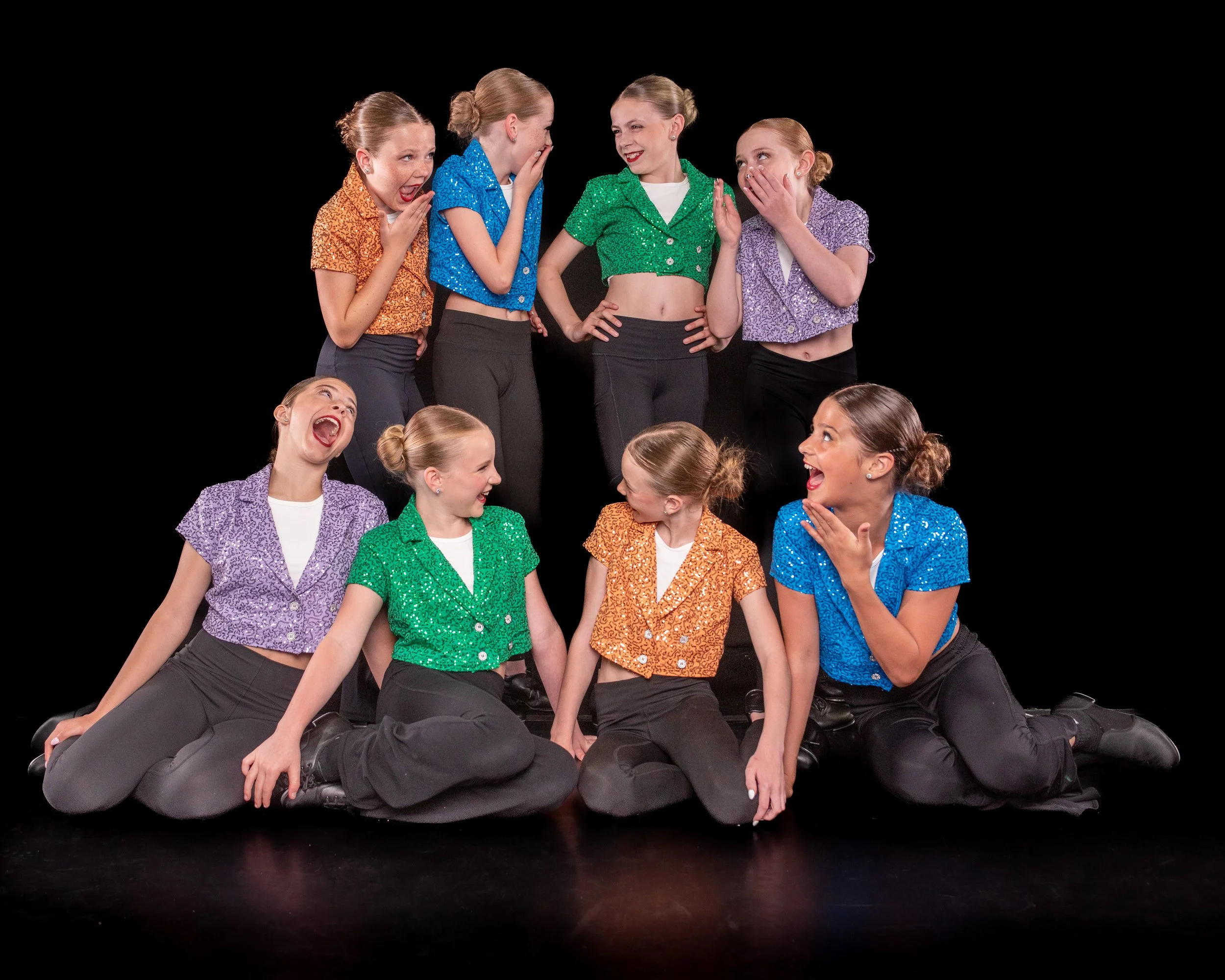 Group of young girls in colorful, sequined dance costumes performing a dance routine on stage with a black background.