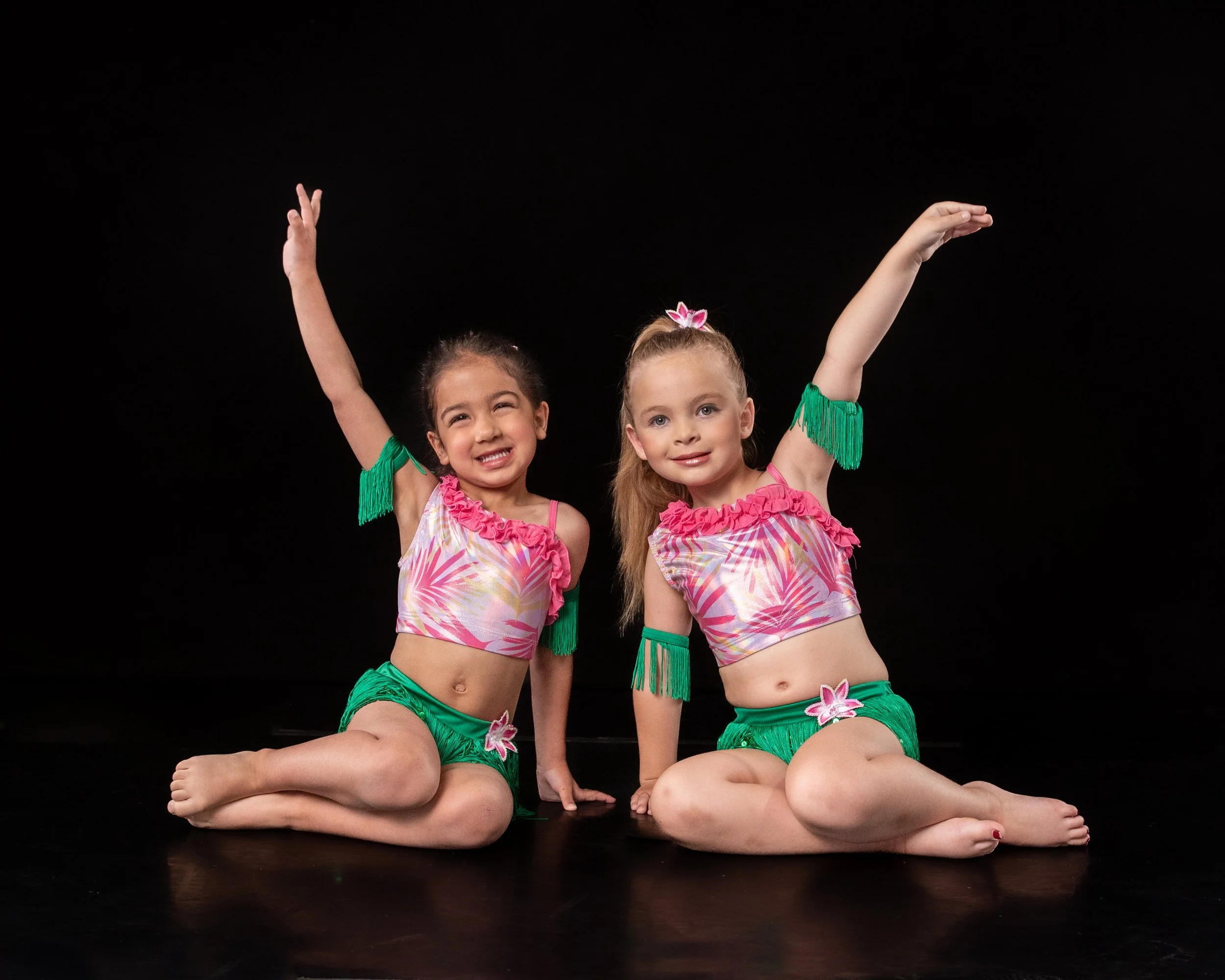 Two young girls dressed in pink and green costumes with floral accents, posing on a black backdrop. One girl is sitting cross-legged and the other is kneeling, both raising one arm above their heads.