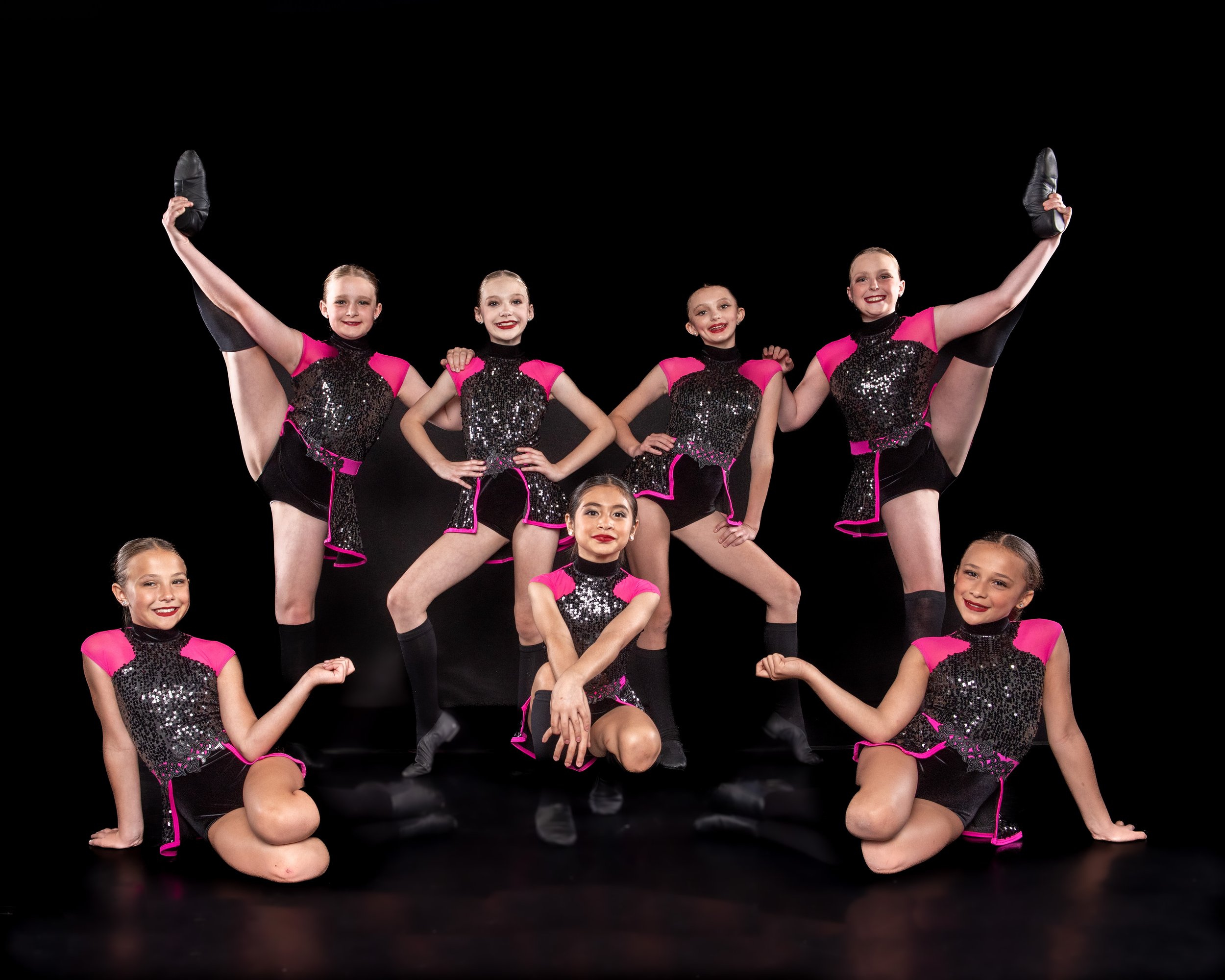 Group of young female dancers in matching pink and black sequined costumes, performing a split pose against a black background.