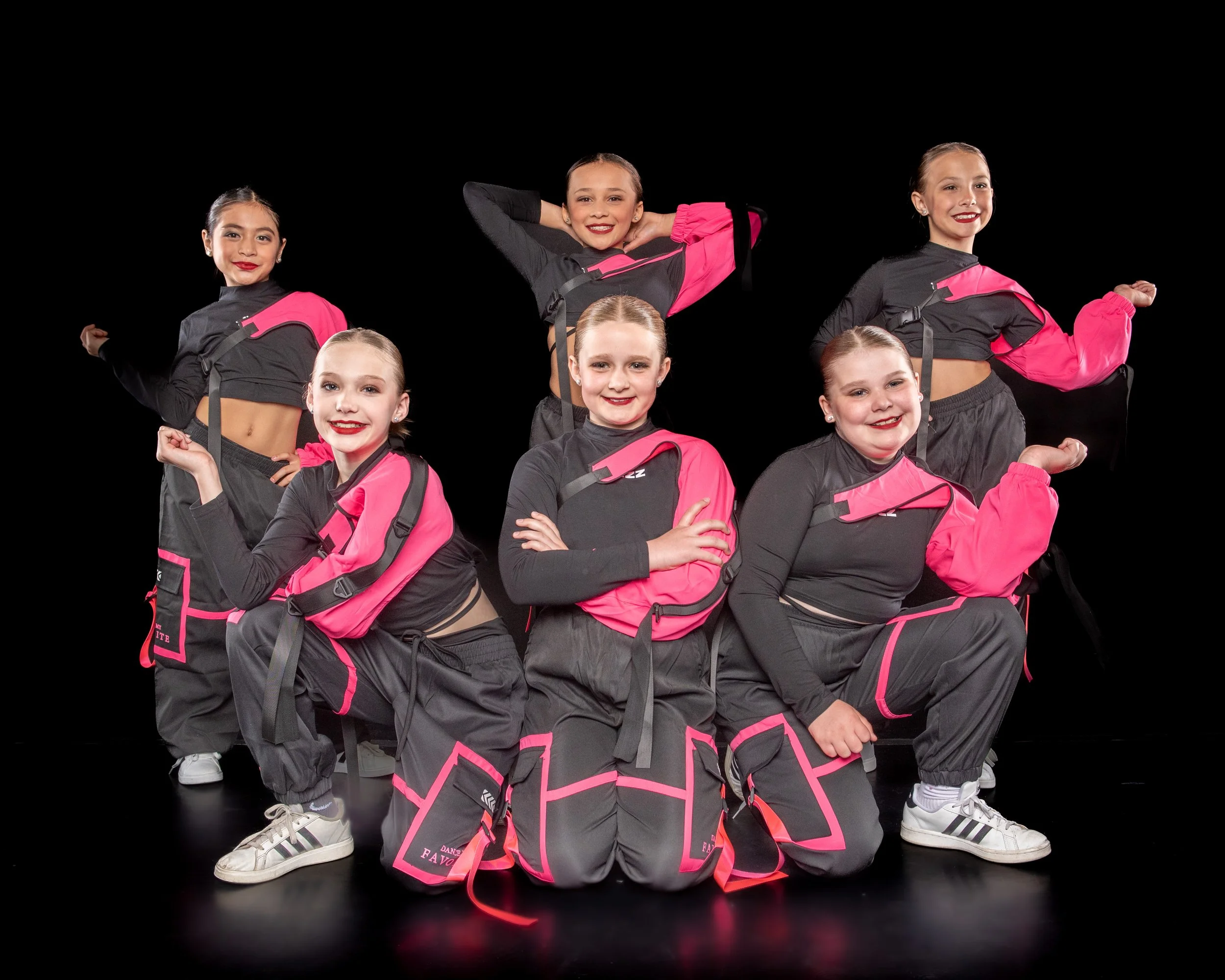 Six young girls in matching black and pink dance outfits, posing confidently against a black background.