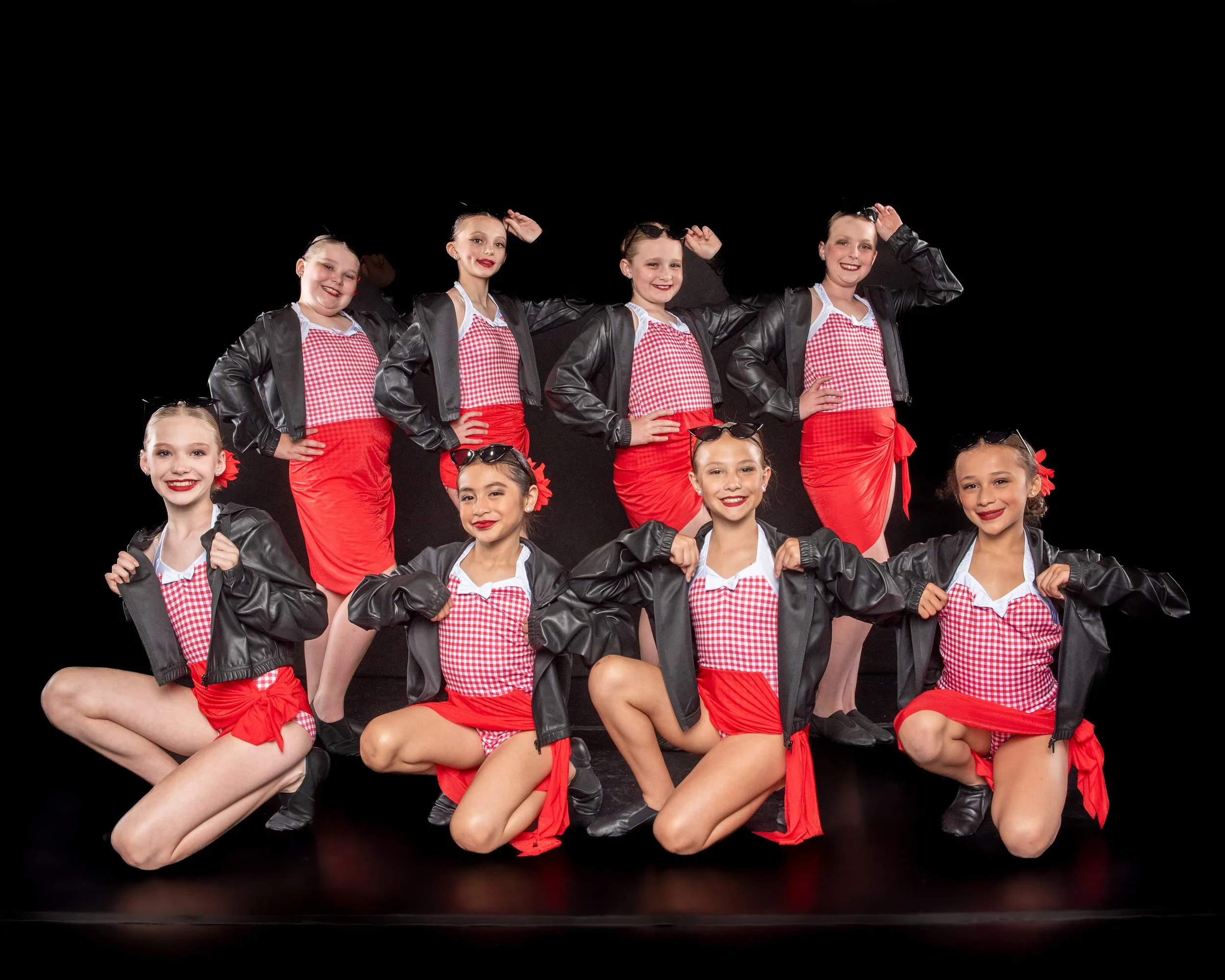 A group of young girls dressed in matching outfits, red skirts, checkered tops, black jackets, and sunglasses, posing on a stage with a black background.