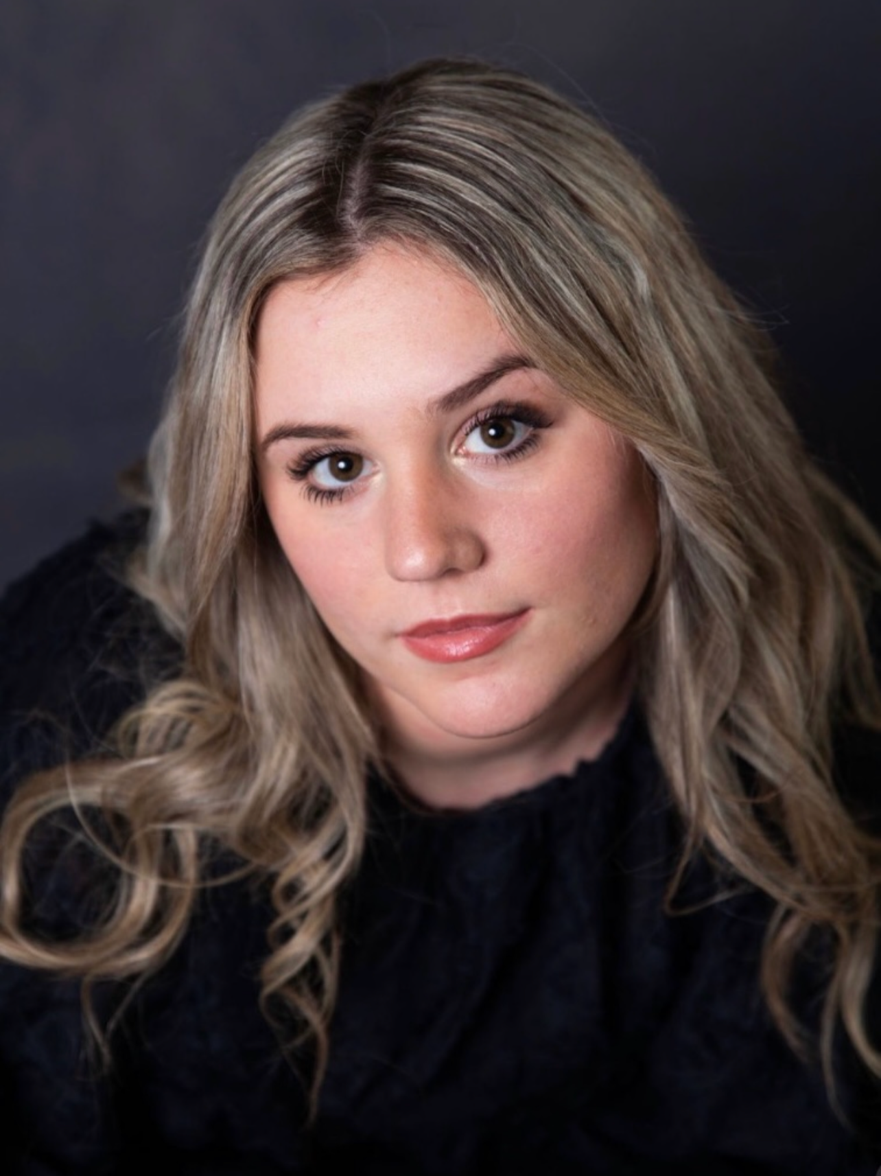 Close-up portrait of a young woman with long wavy blonde hair, wearing a black top, against a dark background.