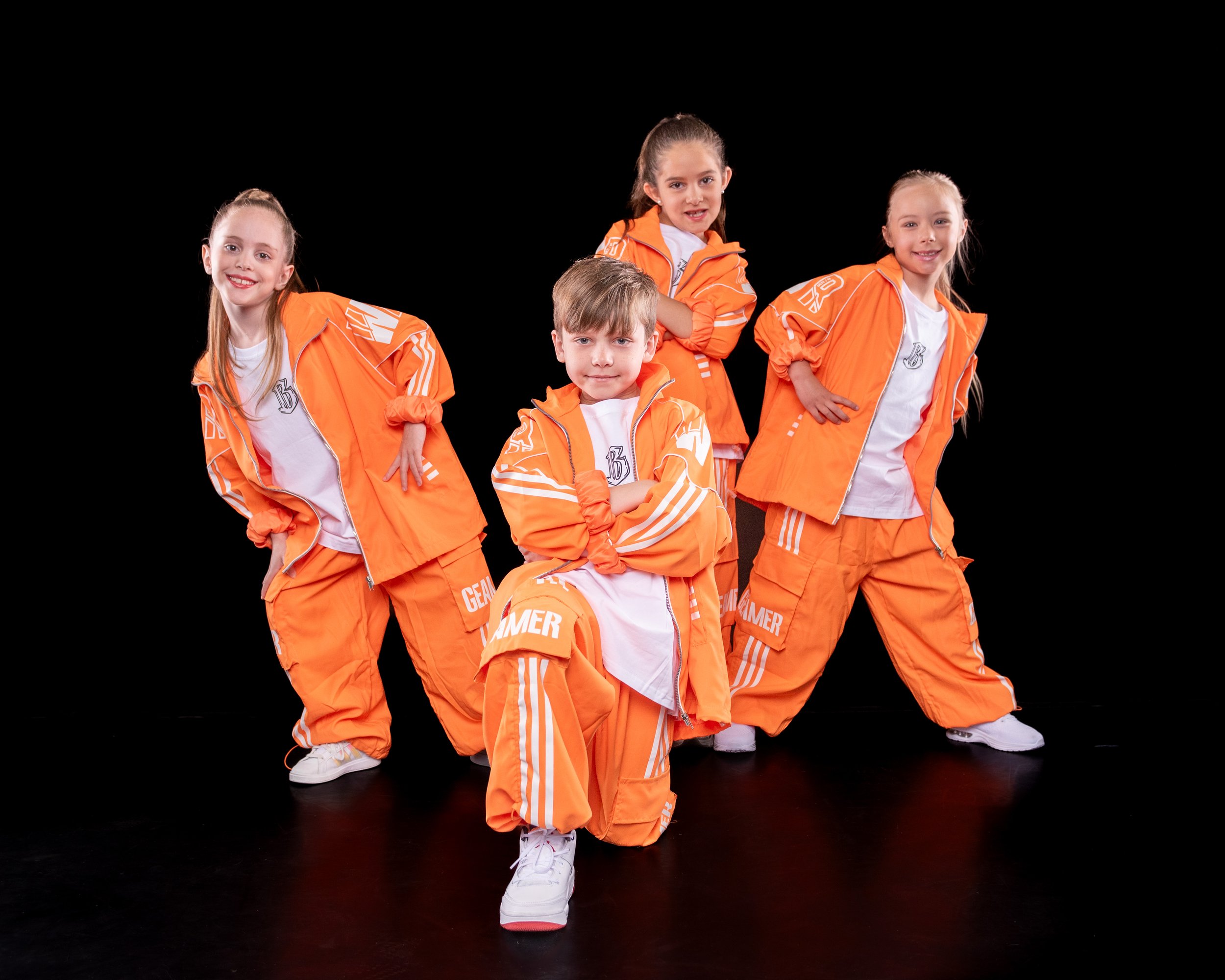 Five children dressed in orange tracksuits are posing together against a black background. They are smiling and looking at the camera, with one child kneeling in front and the others standing behind him.