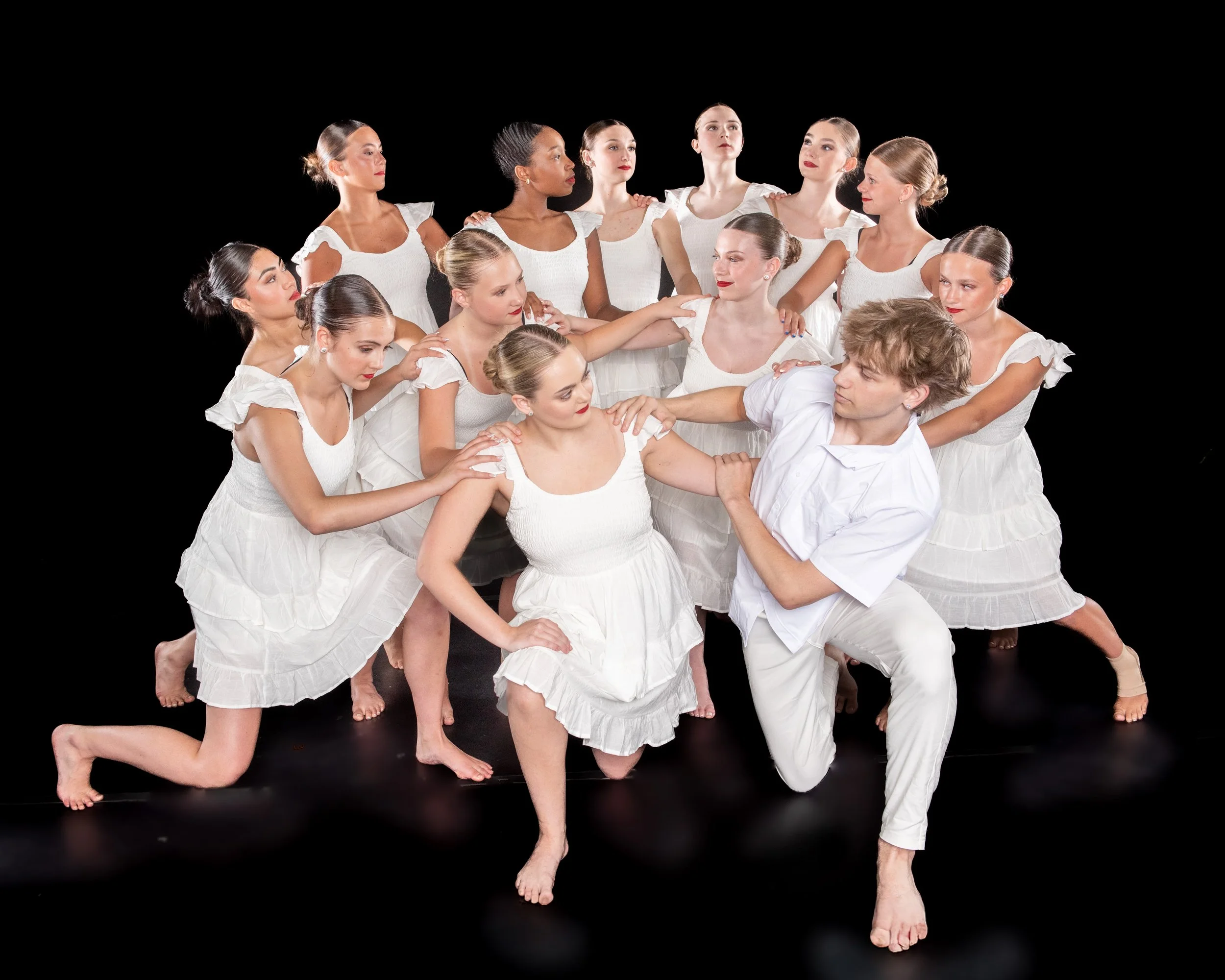 Group of ballet dancers and a male dancer in white costumes performing a dance routine against a black background.