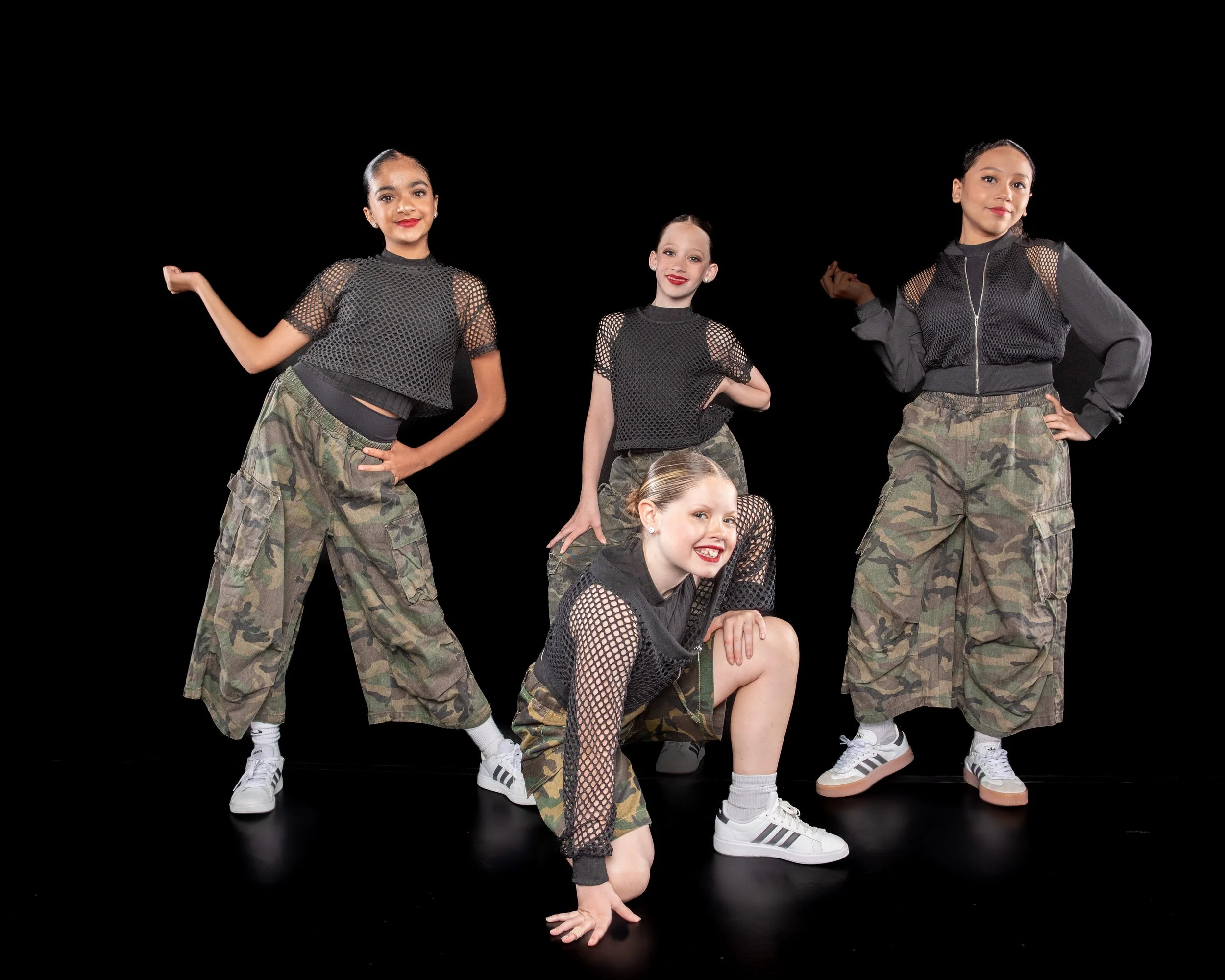 Group of five young girls in athletic outfits with camouflage pants and black mesh tops, posing against a black background, some standing and some crouching, smiling and looking confident.