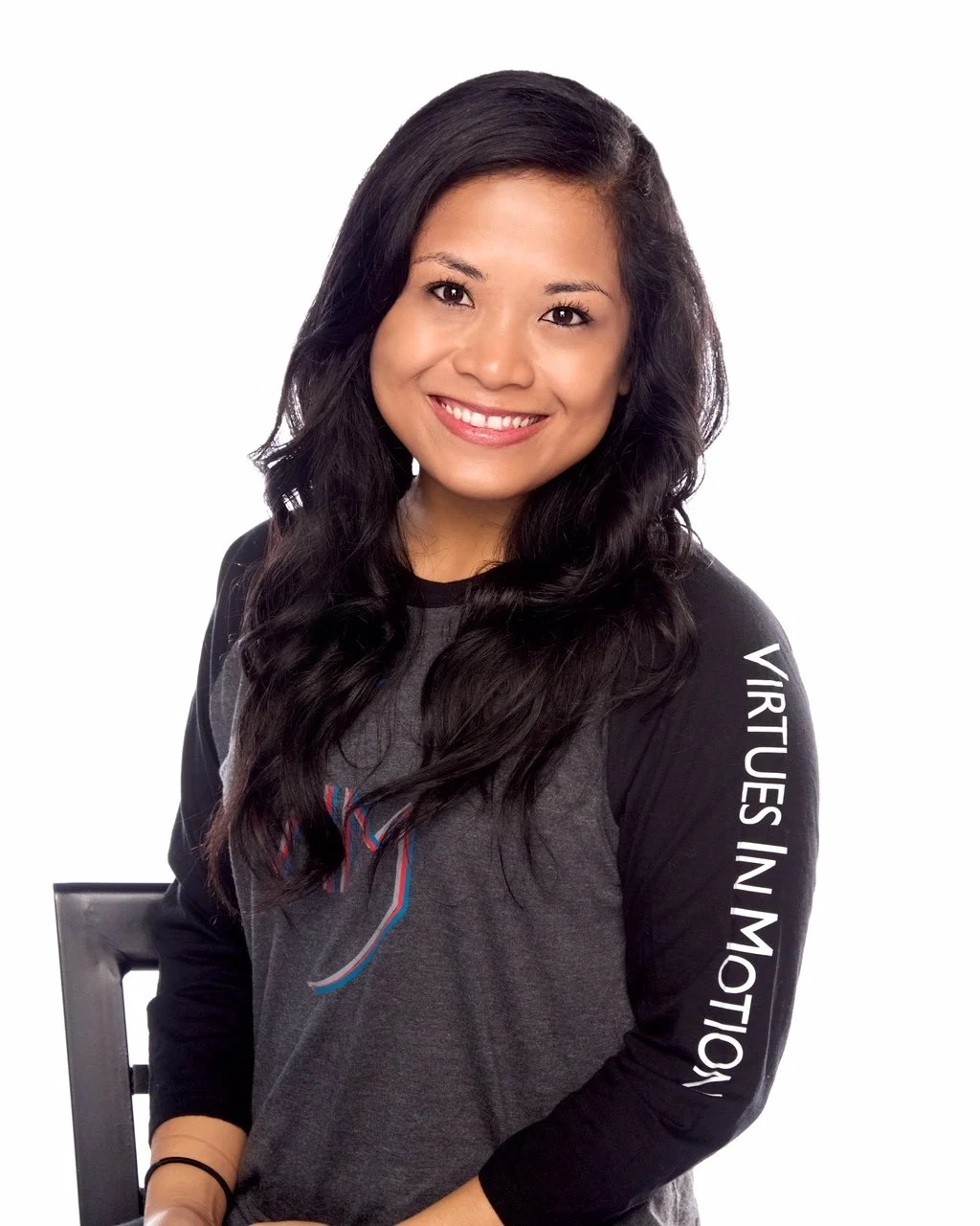 Young woman with long black hair smiling, wearing a gray and black long sleeve shirt with white text on the sleeve and a design on the chest, sitting on a black chair with a white background.
