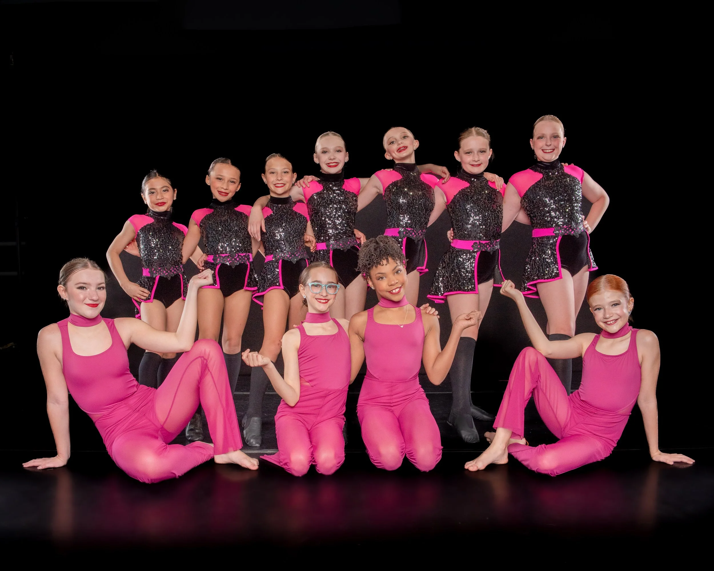 A group of young female dancers in pink and black costumes poses on stage with a black background.