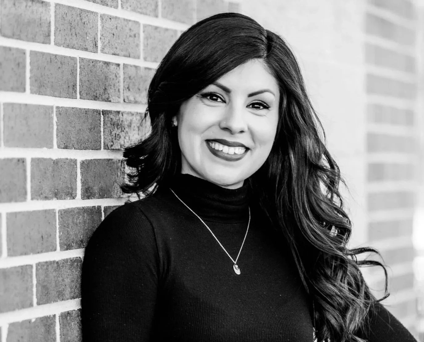 Black and white portrait of a woman with long wavy hair, wearing a black turtleneck and a pendant necklace, smiling, standing against a brick wall.