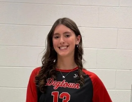 Young woman with long brown hair wearing a black and red volleyball jersey with the number 13 and 'Dig Town' written on it, standing against a white brick wall and smiling.