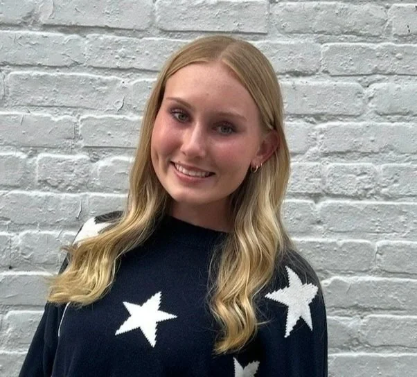 A young woman with long blonde hair smiling, wearing a black sweater with white star patterns, standing in front of a white brick wall.