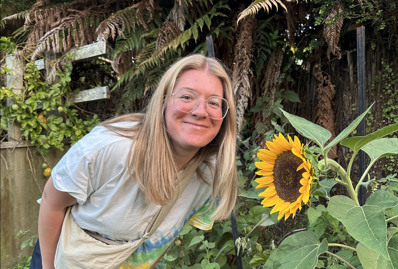 Photo of Emily with a rainbow tie dye shirt smiling next to a large sunflower.