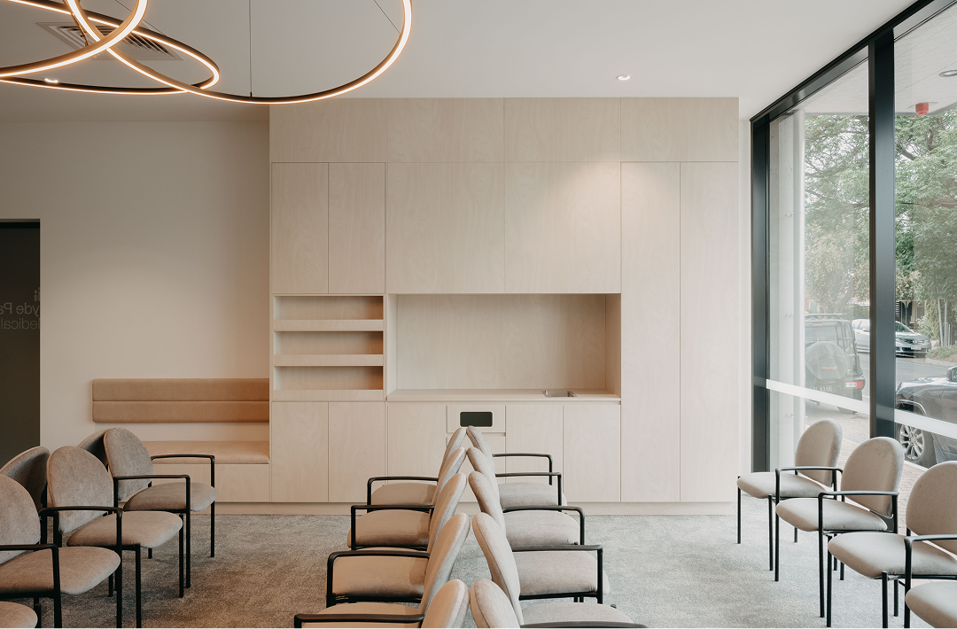 Hyde Park Medical Clinic waiting room featuring soft neutral tones, light timber joinery, upholstered chairs with black frames, and a sculptural circular pendant light, with floor-to-ceiling windows overlooking a tree-lined street.