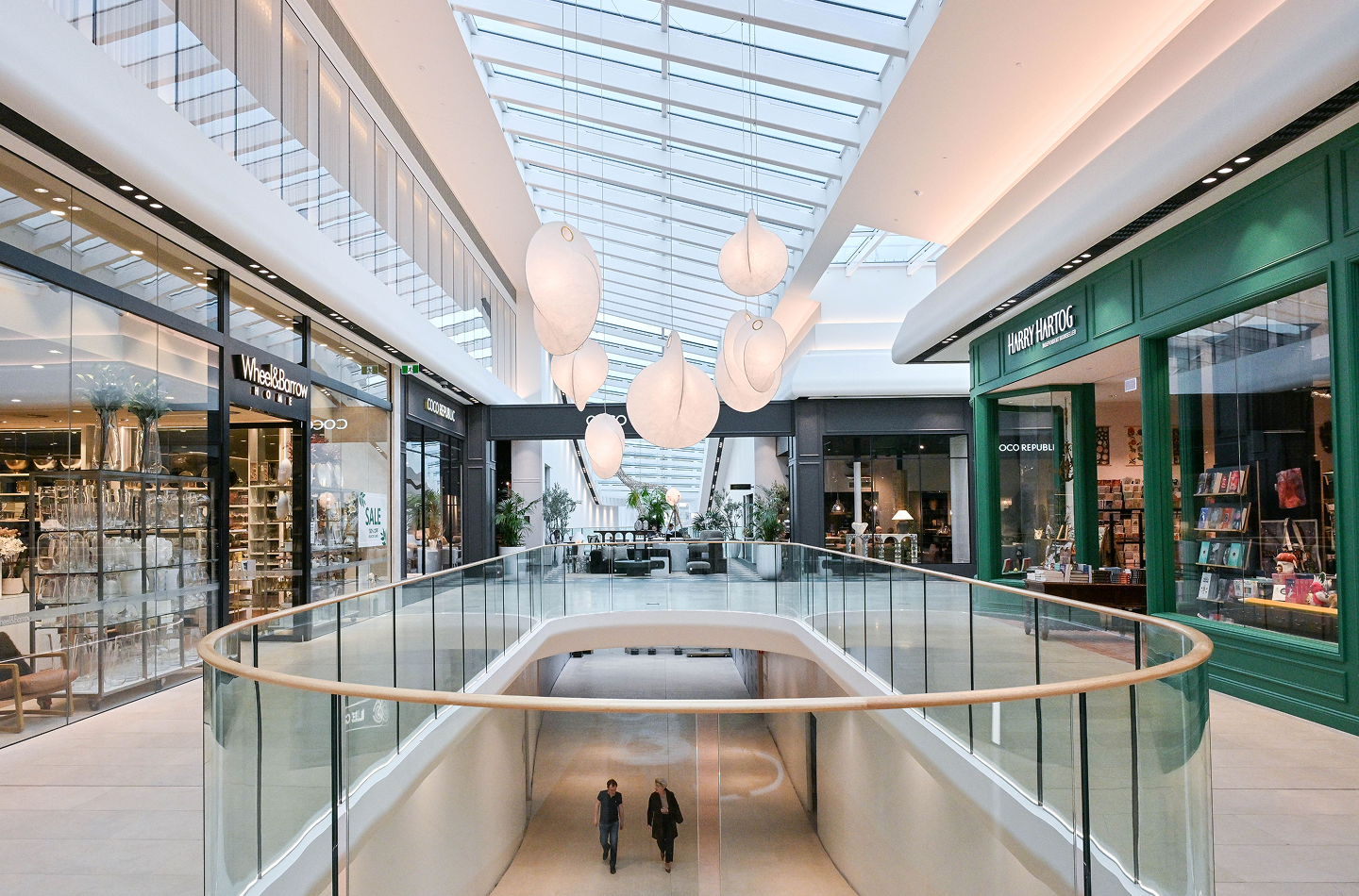 Burnside Village upper level retail precinct with a full-length glass skylight, sculptural white pendant lights, curved glass balustrade with timber handrail, and premium retailers including Wheel & Barrow, Coco Republic, and Harry Hartog.