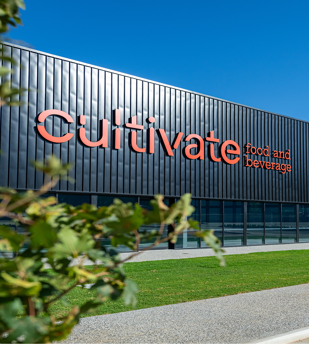 Exterior of Cultivate Food and Beverage facility featuring dark ribbed metal cladding with bold orange signage, set against a clear blue sky with green landscaping in the foreground.