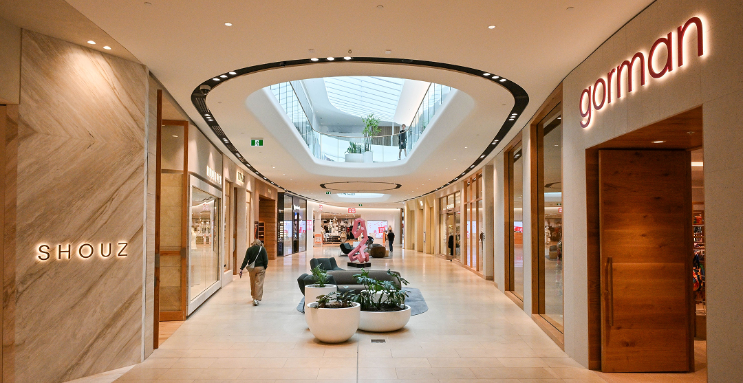 Burnside Village shopping centre retail laneway featuring warm stone and timber finishes, an oval skylight, potted greenery, and retailers including Shouz and Gorman.