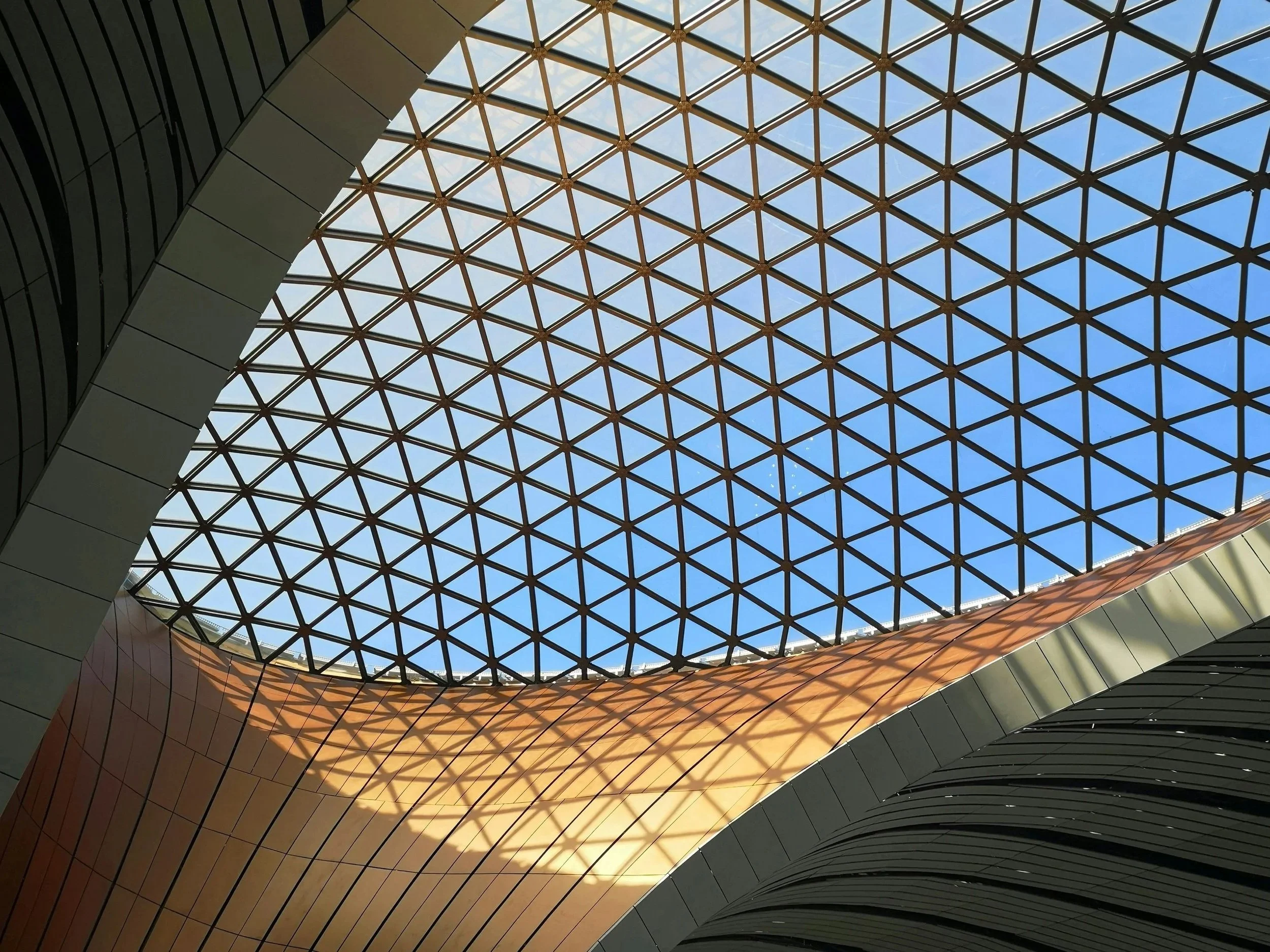 Upward view of a geometric triangulated glass roof structure with dark steel framing, casting warm shadow patterns onto curved copper-toned cladding beneath a clear blue sky.