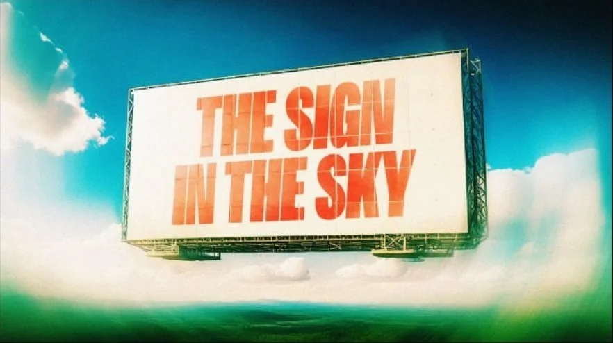 Billboard with the bold text, 'THE SIGN IN THE SKY,' floating in the sky above the ocean with a partly cloudy background.