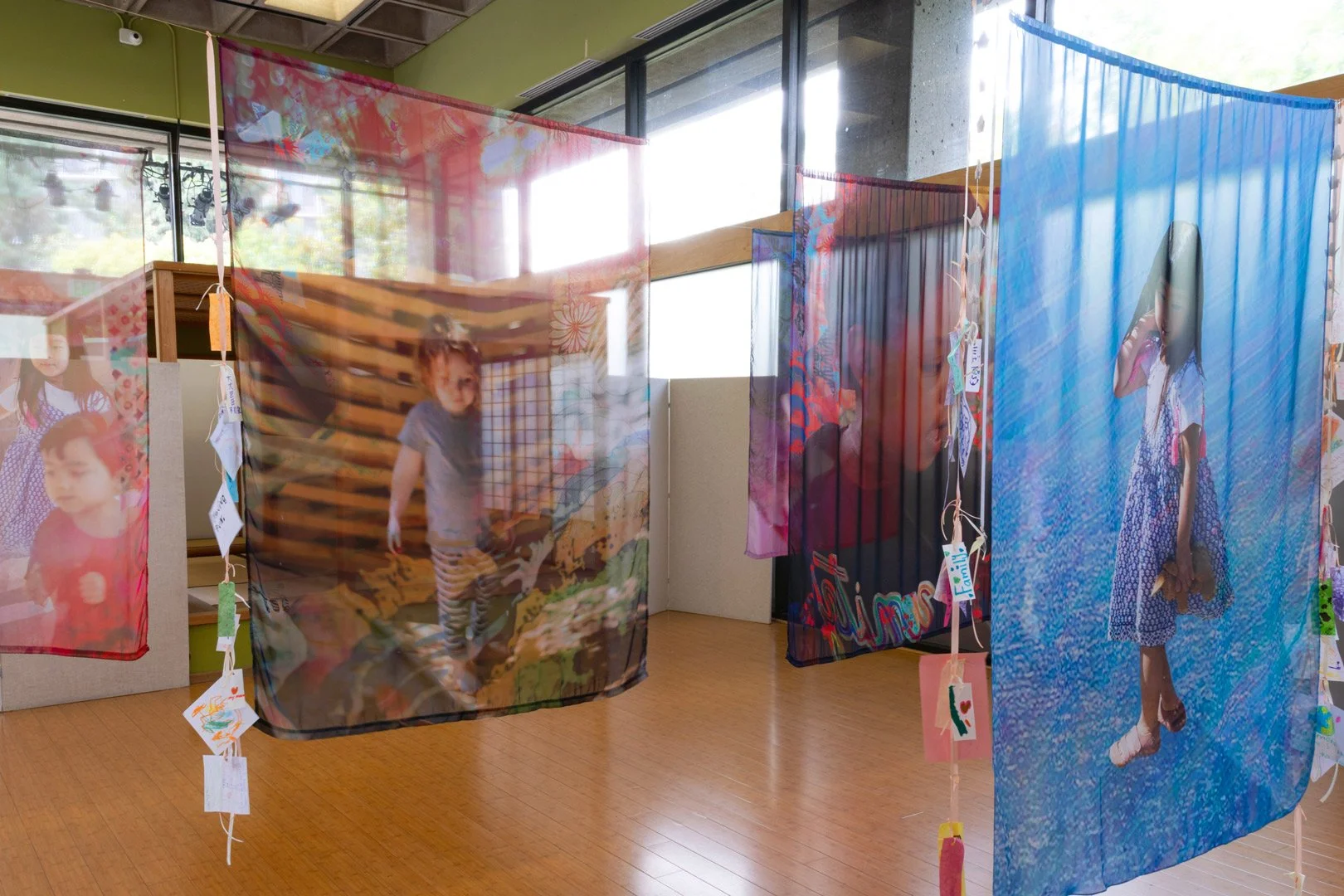 Colorful hanging fabric art with printed photographs of children and handwritten notes, displayed in an indoor gallery with large windows and wooden floors.