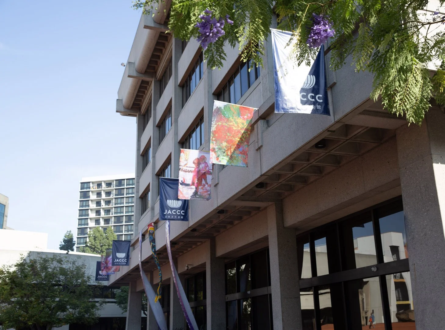 Exterior of a modern building with flags and colorful banners hanging from the balconies. The building has multiple floors and large windows, with trees and a clear sky in the background.