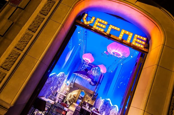 Vitrina de tienda con decoración temática de globos aerostáticos rosas y un letrero que dice 'VERNE', iluminado sobre un fondo azul.