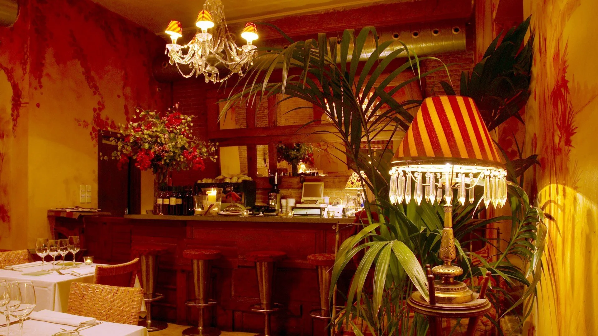 Interior del restaurante elegante con iluminación cálida, mesas con manteles blancos y copas de vino, un candelabro colgante, una lámpara vintage y decoración floral exuberante.