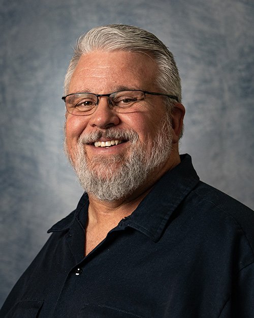 Smiling older man with gray hair, glasses, wearing a blue t-shirt outdoors with green trees and a clear sky in background.