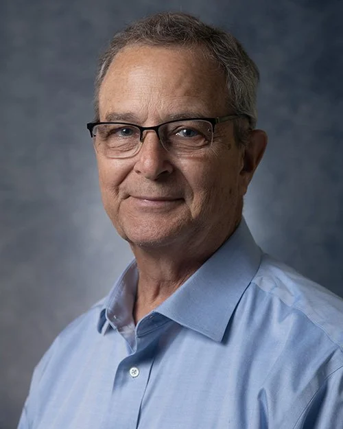 Portrait of a middle-aged man in a dark suit, white shirt, and blue tie, smiling against a plain white background.