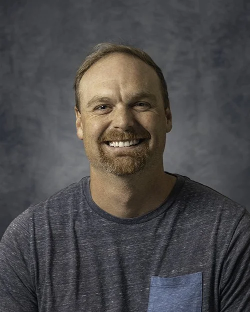 A smiling man with a beard and short hair standing outdoors on a sunny day, wearing a dark gray t-shirt, with trees and a parking lot in the background.