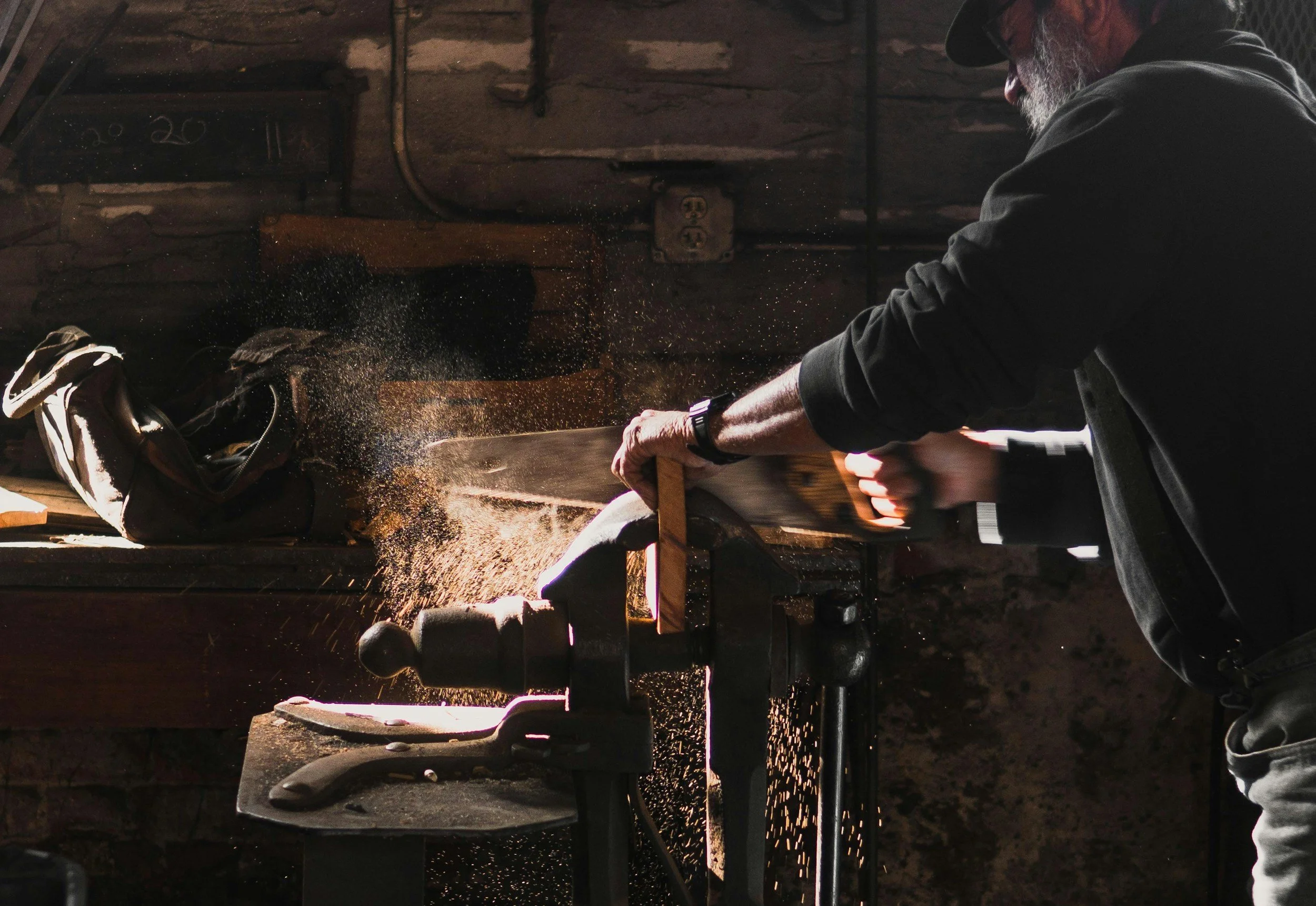 Bearded man sawing lumber with a handsaw in a woodworking shop with a light beam shining through the wood dust.