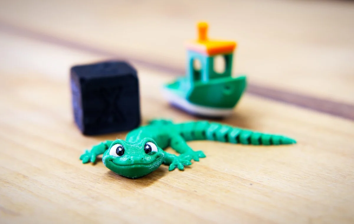 A green 3d printed gecko on a wooden bench top with a 3d printed black cube and a benchy in the background.