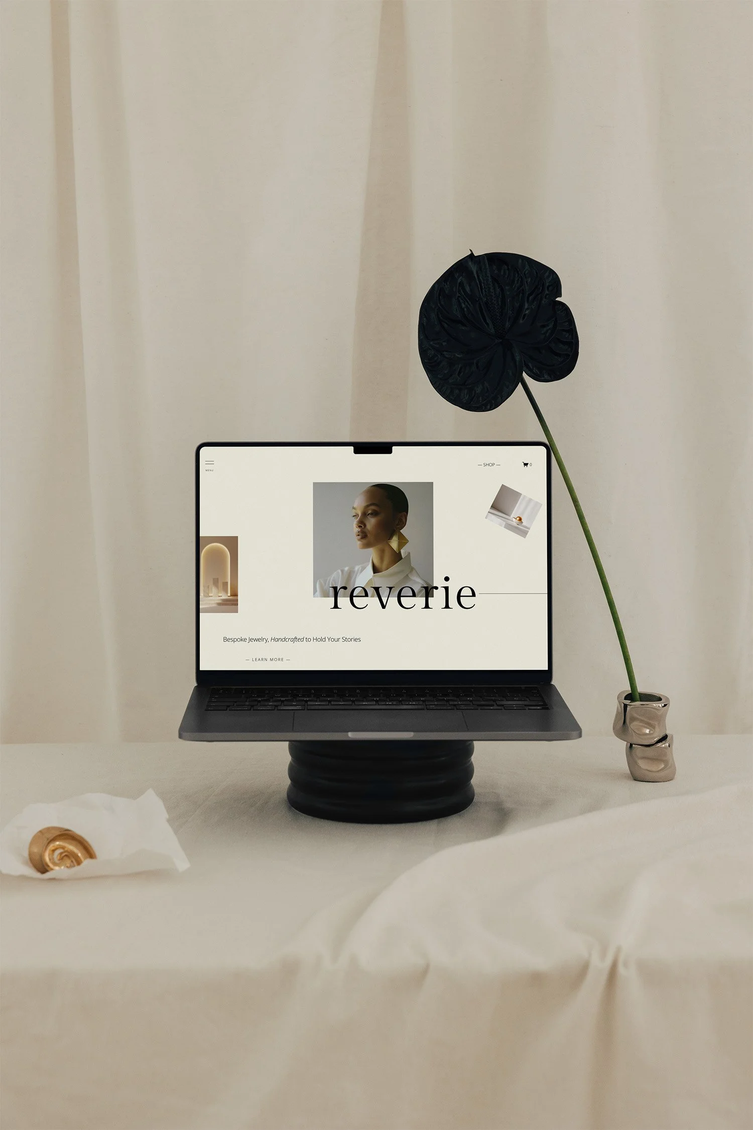 A laptop displaying a jewellery website called Reverie, placed on a black ceramic stand on a beige table. To the right, there's a small vase with a black flower. On the left, there is a golden shell on a tissue.