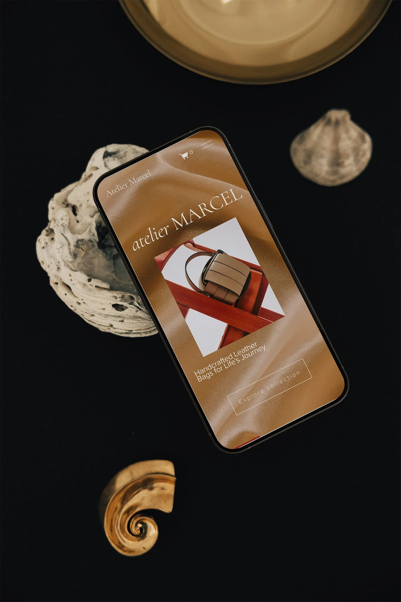 Smartphone displaying the website of atelier MARCÉL, a handbag brand, placed on a black surface along with seashells and a gold spiral ornament.
