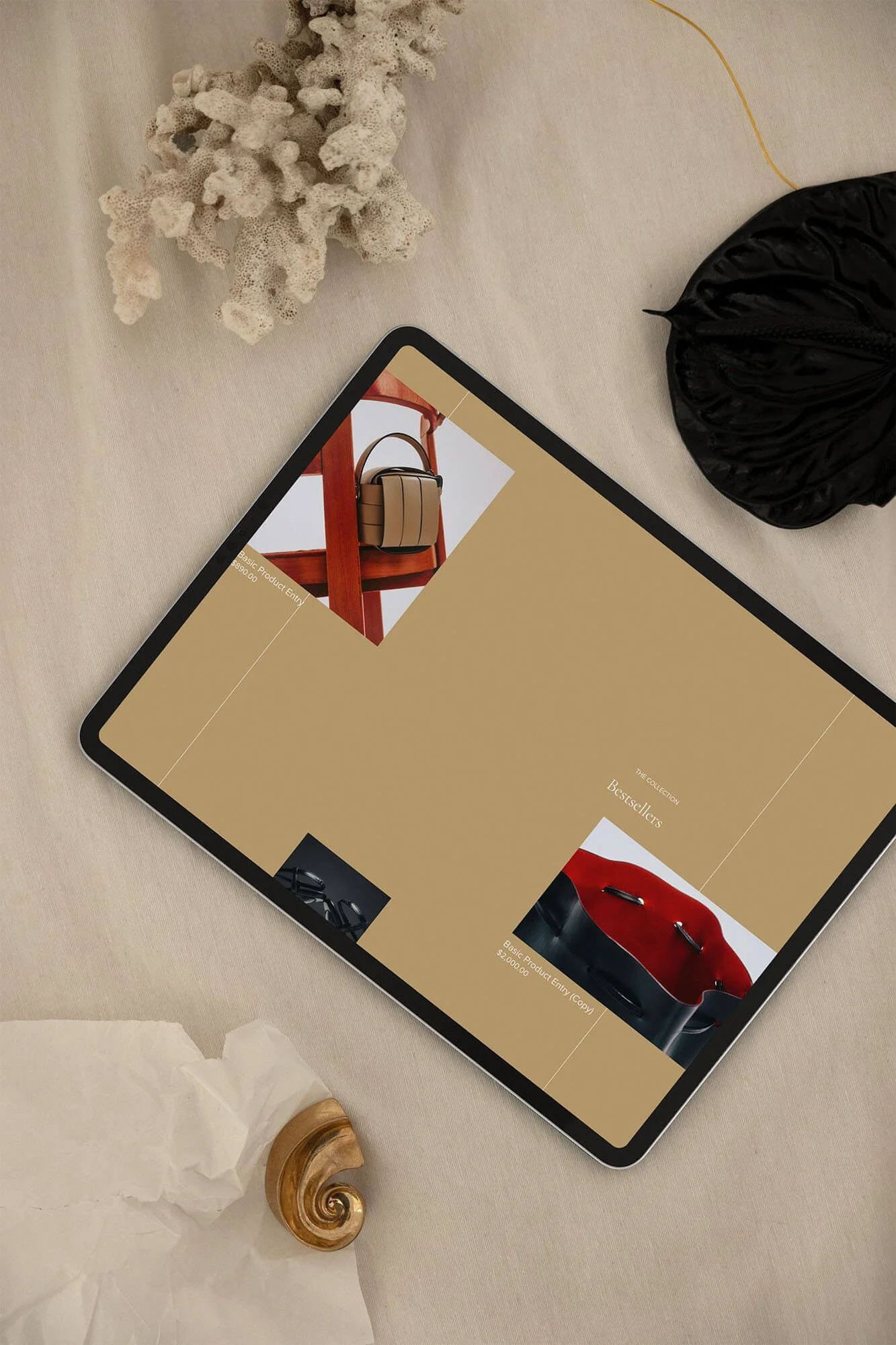 Tablet displaying a website featuring handbags, surrounded by decorative objects including a piece of coral, a black pouch, and a seashell.