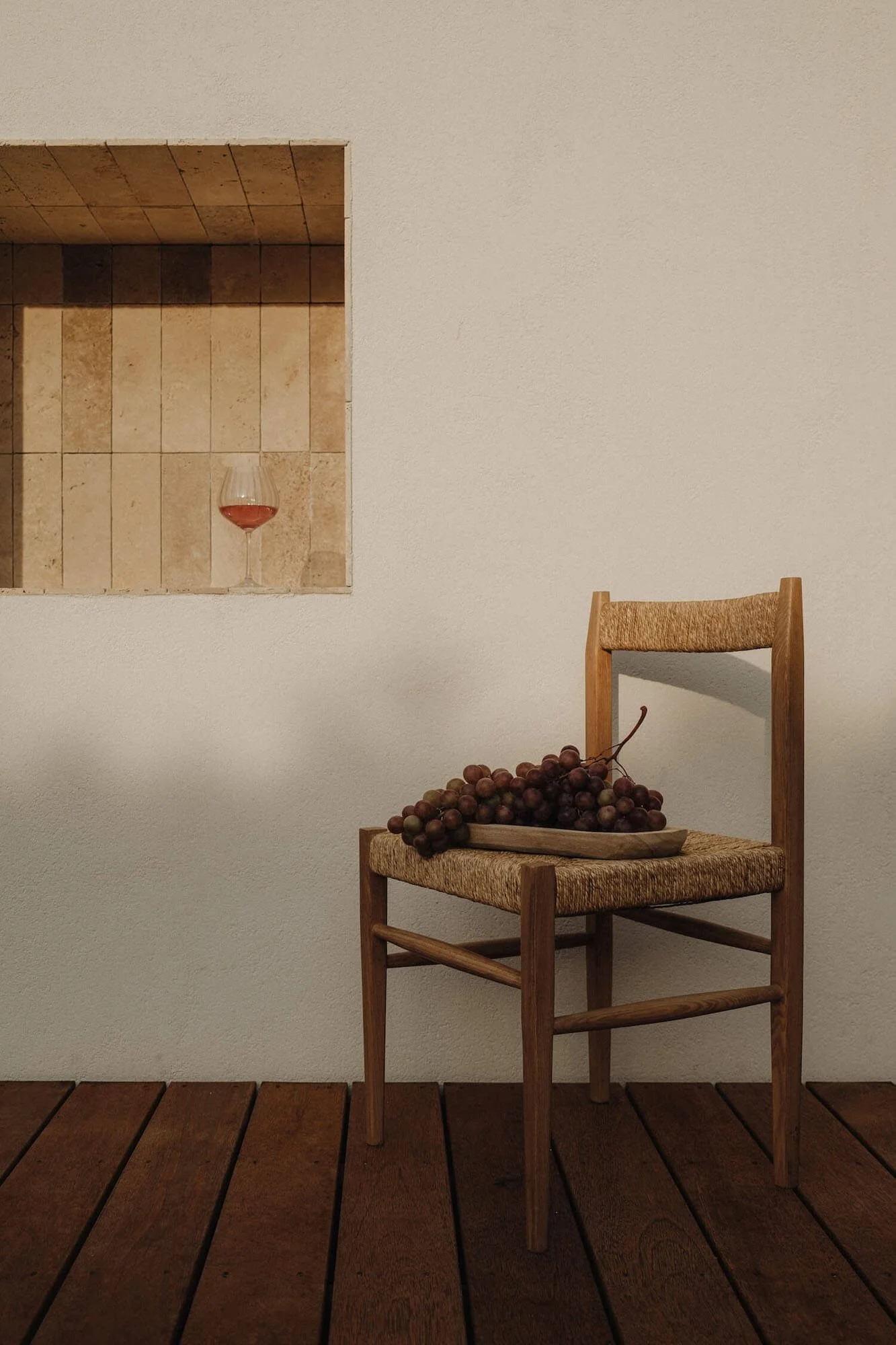 A wooden chair with a bunch of grapes on it, placed on a wooden floor, with a small wall opening in the background containing a wine glass filled with red wine.