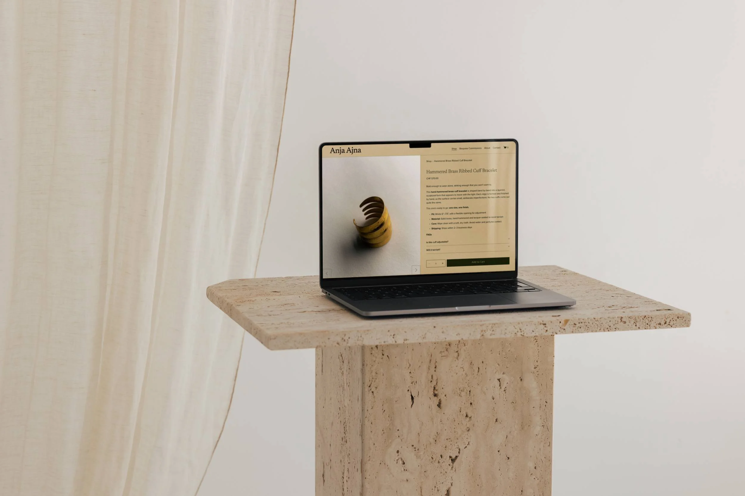 Laptop on a travertine stone pedestal against sheer curtains, displaying the Anja Ajna website product page for a Hammered Brass Ribbed Cuff Bracelet