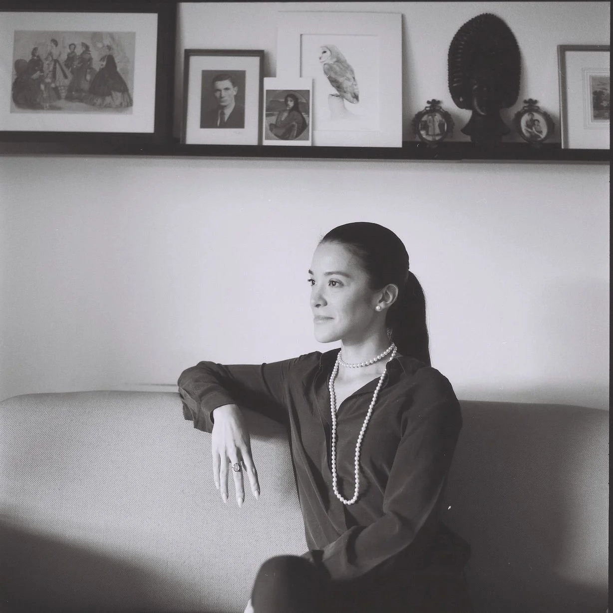 Rebecca Giger, female website designer, hair tied back, wearing a pearl necklace and earrings, a dark blouse, sitting on a sofa with her arm resting on the back, looking to her left. Behind her is a a shelf displaying framed photographs and artwork.