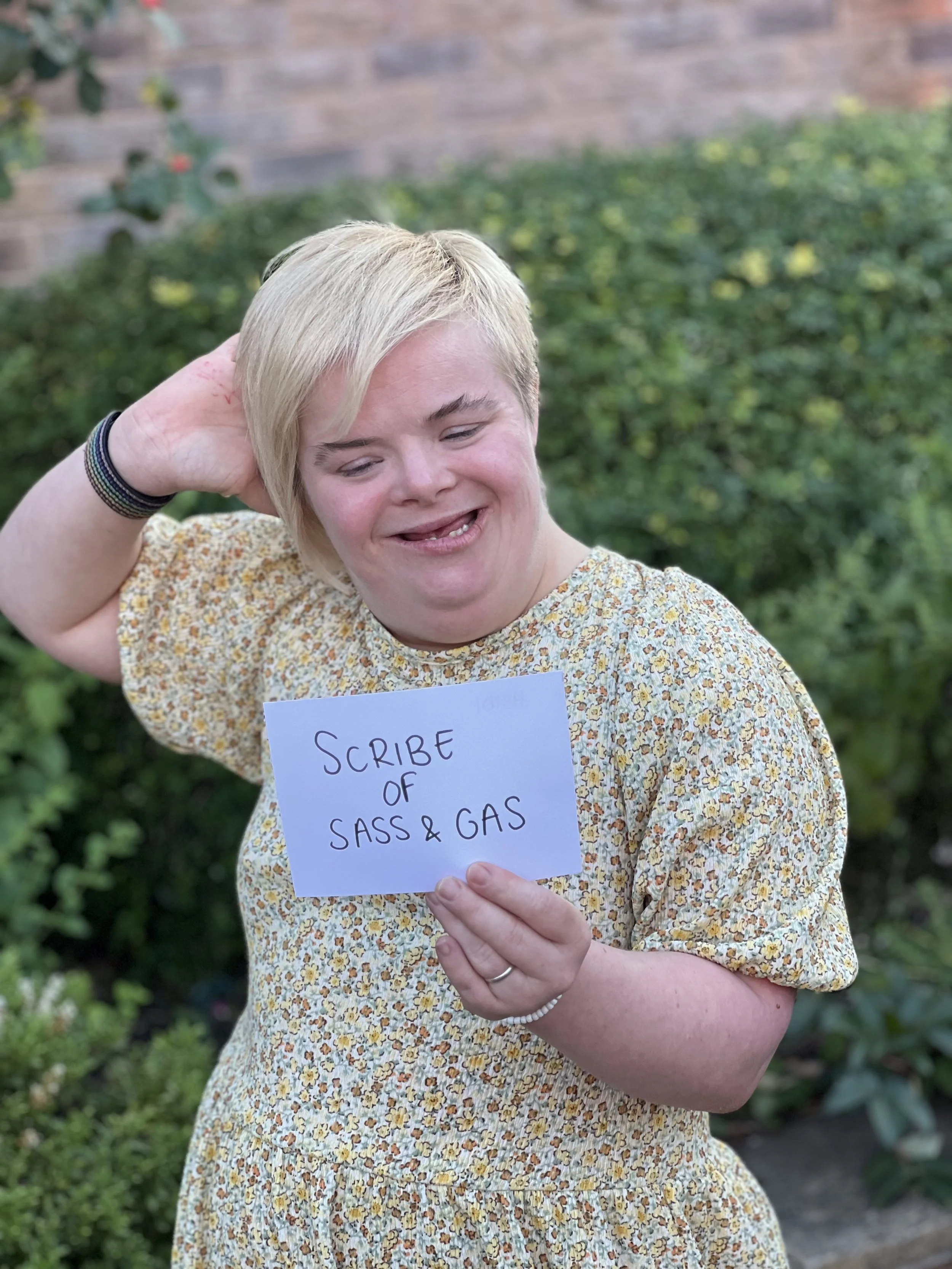A photo of heidi, smiling at the camera with her hand behind her head. Heidi is holding a sign that says "Scribe of Sass & Gas"