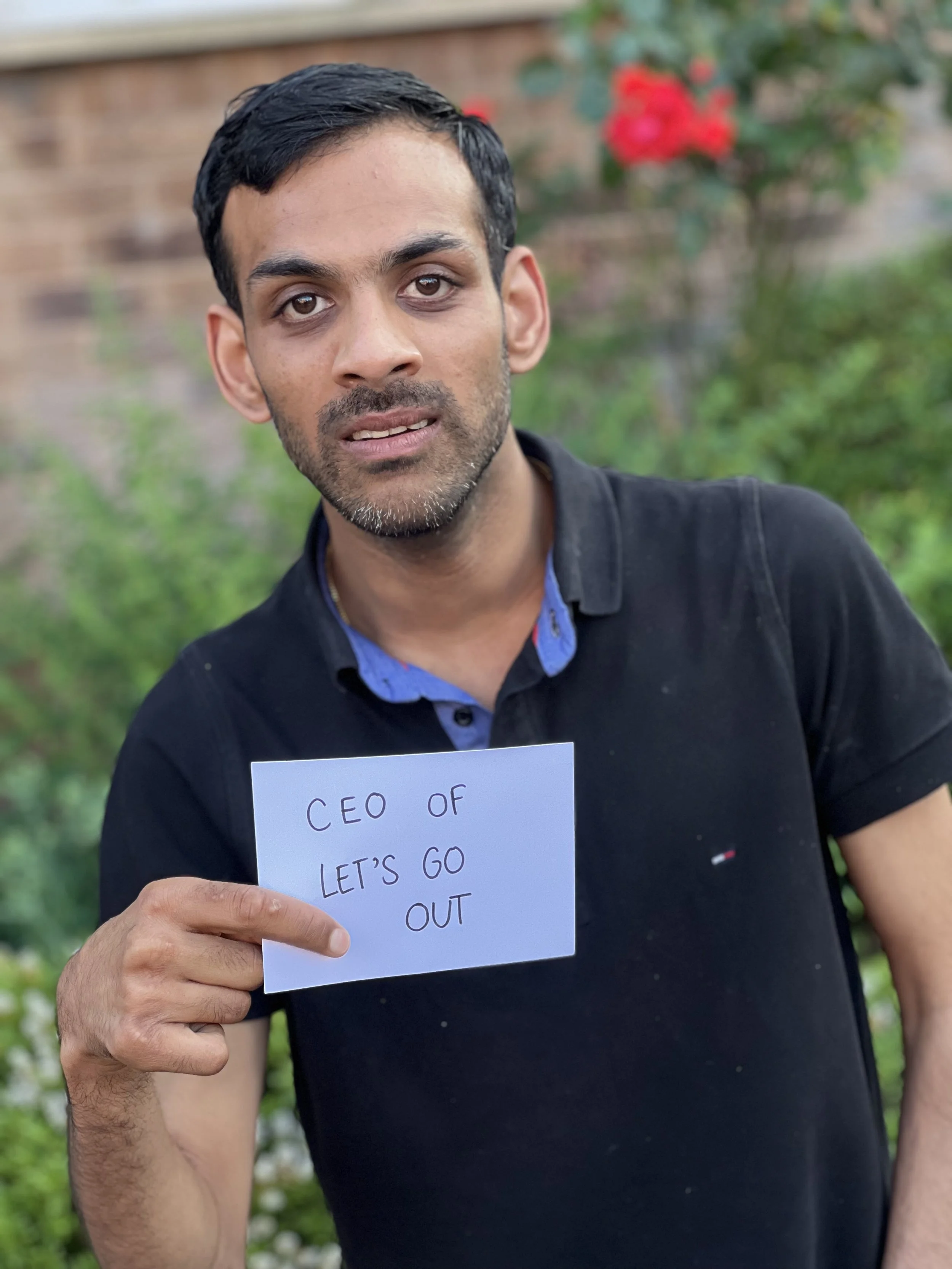 A photo of Prash standing outside. Prash is looking at the camera and holding a sign that says "CEO of Let's Go Out"