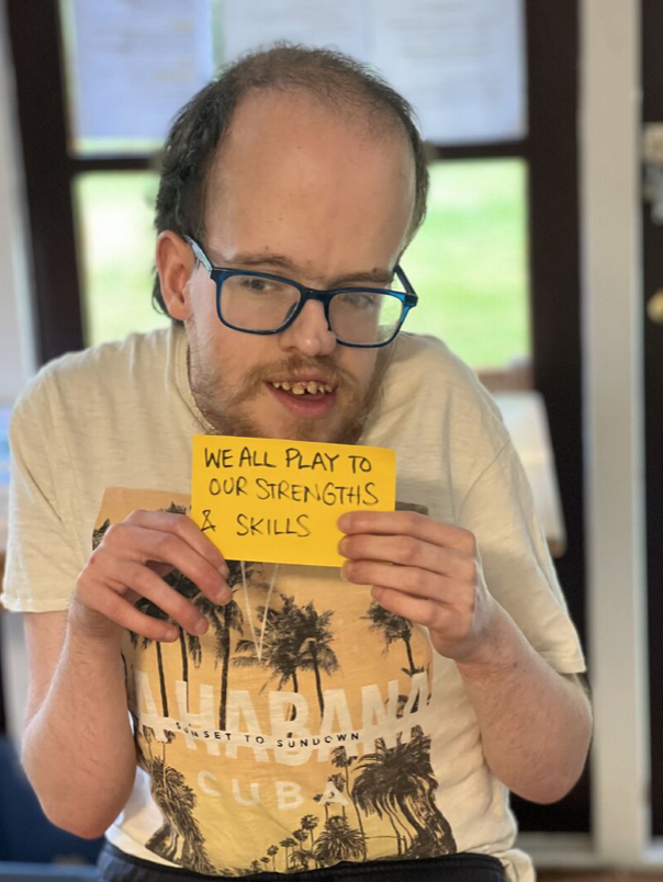 A photo of Thomas wearing blue glasses. Thomas is smiling and holding a sign that says "We All Play To Our Strengths & Skills"